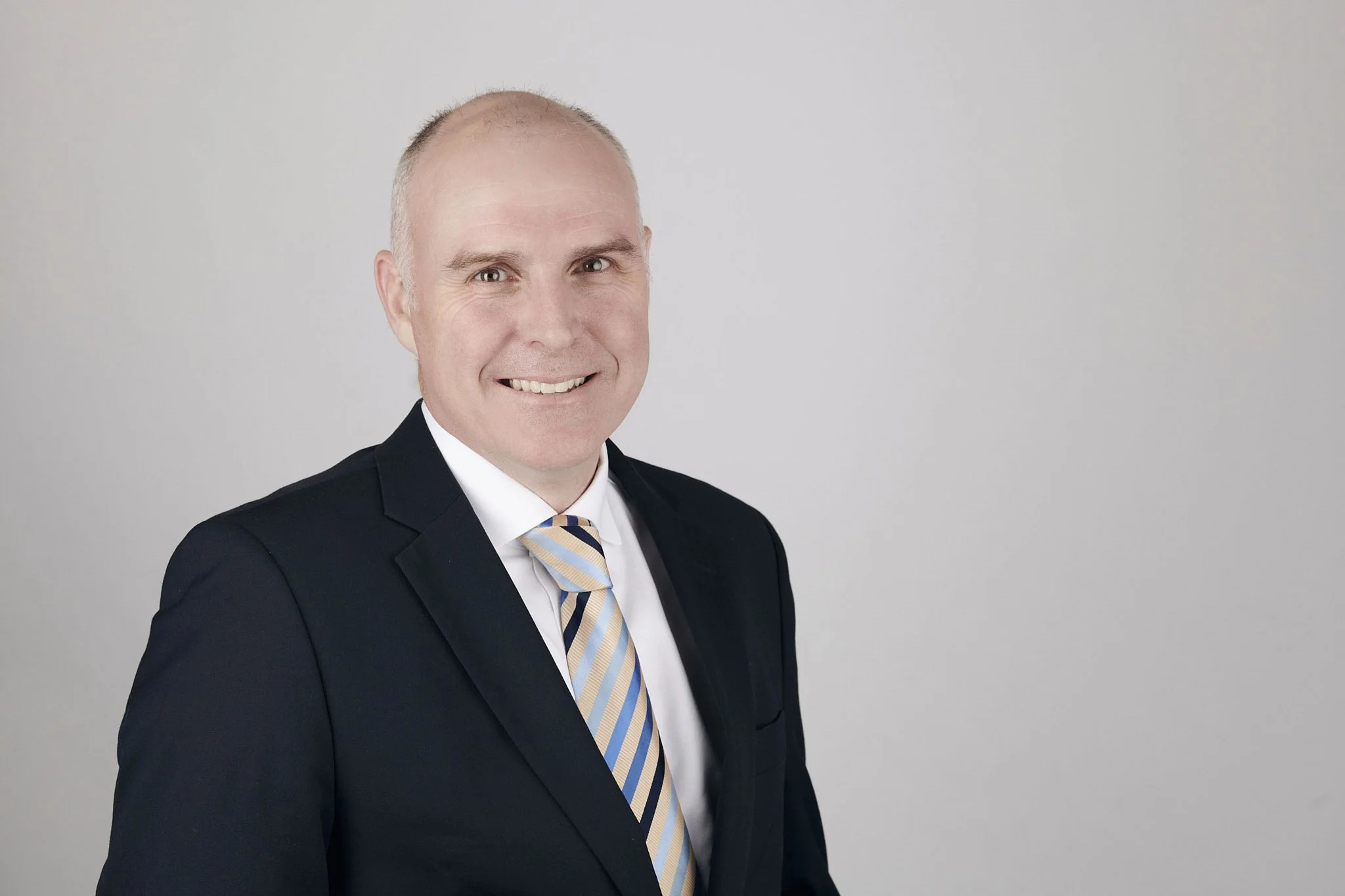 A smiling man in a black suit, white shirt, and striped tie posing against a plain white background.