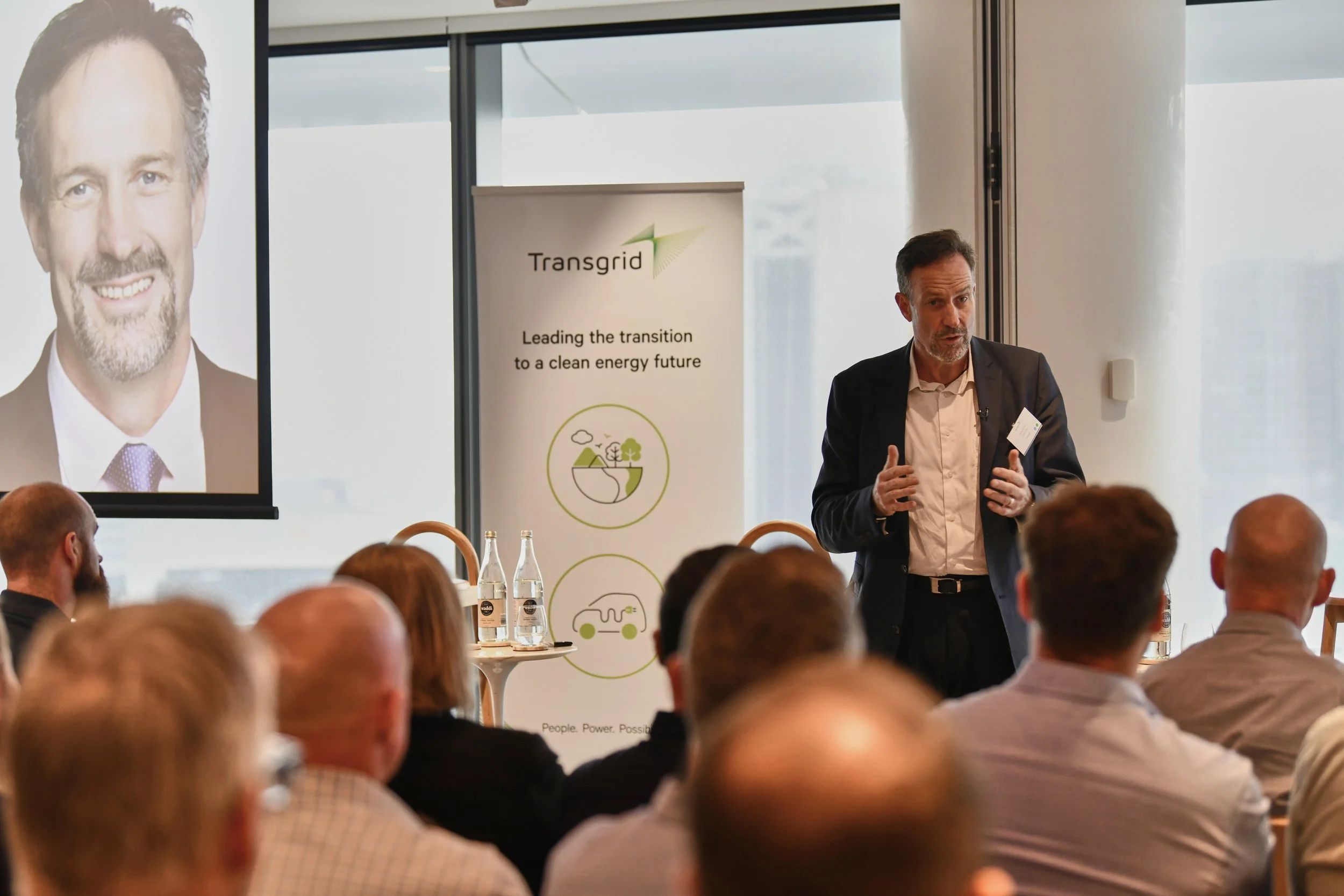 A man presenting at a conference, standing in front of an audience with a large screen displaying a man's portrait to his left and a banner with the logo 'Transgrid' behind him, emphasizing clean energy transition.