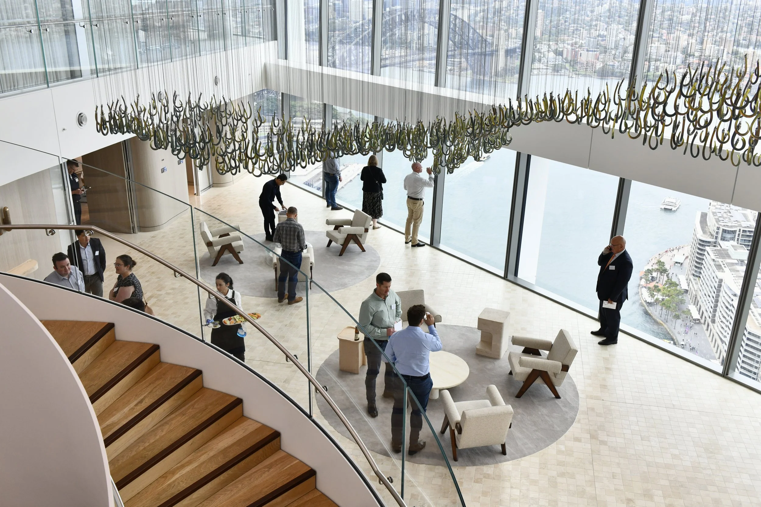People gathered in a modern, high-rise lobby with large glass windows overlooking a city and water, featuring a contemporary chandelier hanging from the ceiling and a staircase with wooden steps.