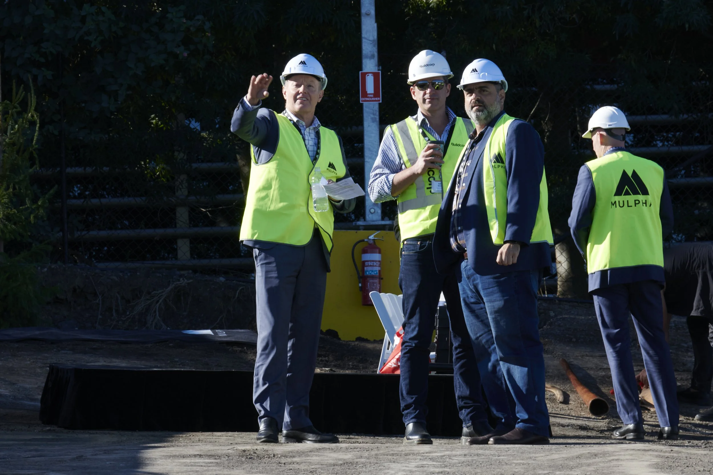 Four men in business suits wearing safety vests and helmets, standing outdoors at a construction site, one is waving, and another is holding a phone.