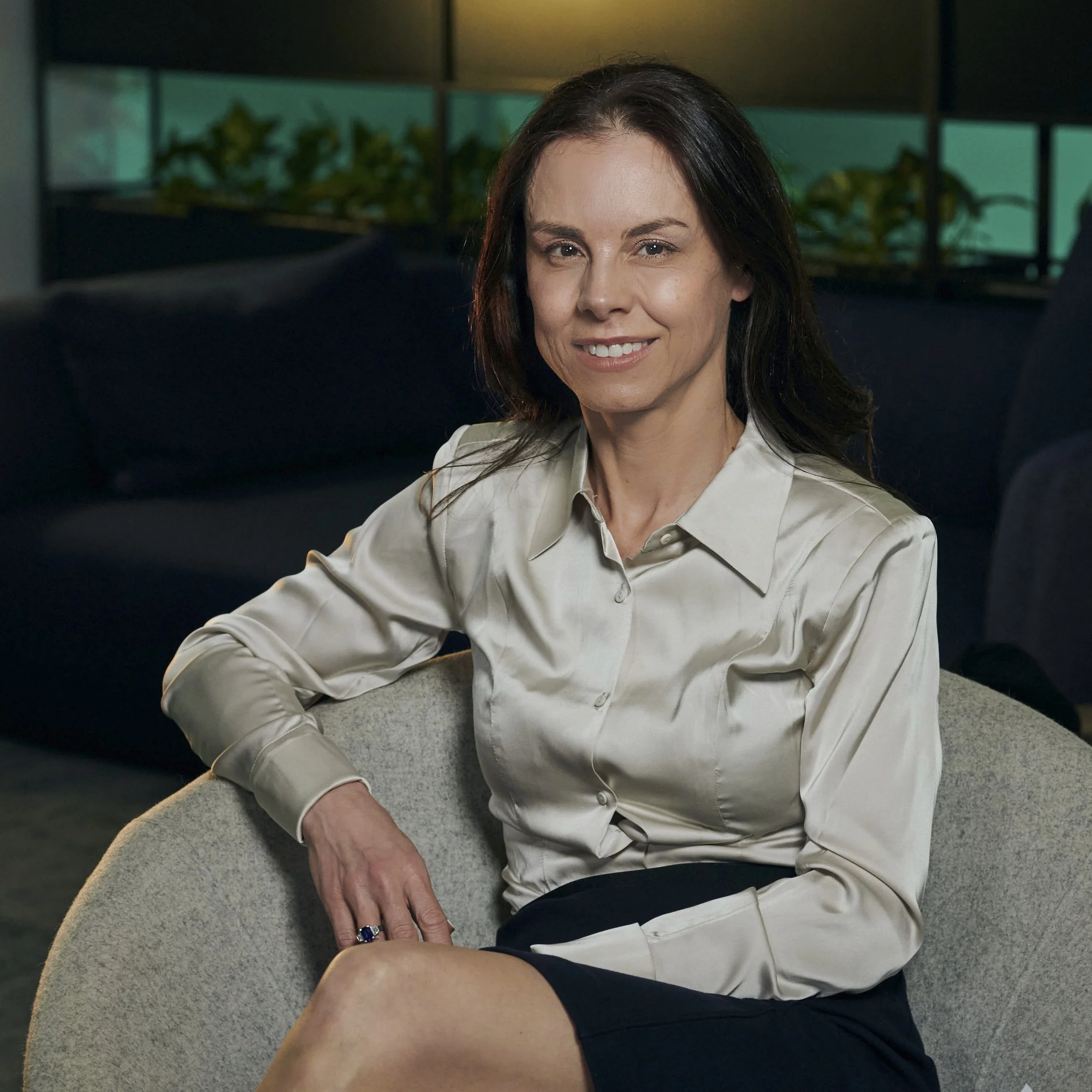 A woman with dark brown hair, wearing a cream-colored satin blouse and a dark skirt, sitting on a light grey chair, smiling at the camera in a modern office or lounge setting.