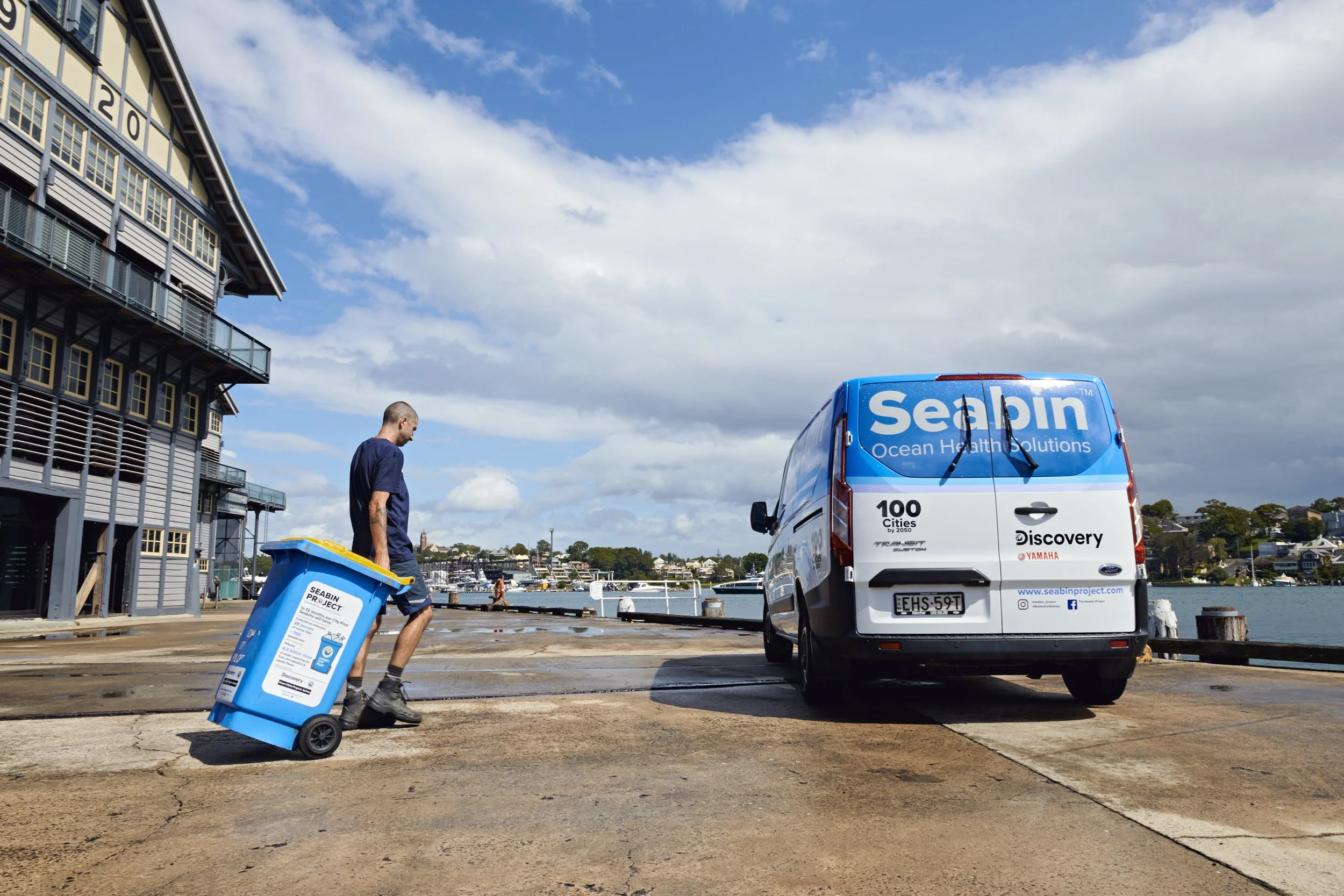 A man pulling a blue recycling bin labeled 'Seabin Project' along a dock near water, with a white van marked for 'Seabin Ocean Health Solutions' parked nearby.