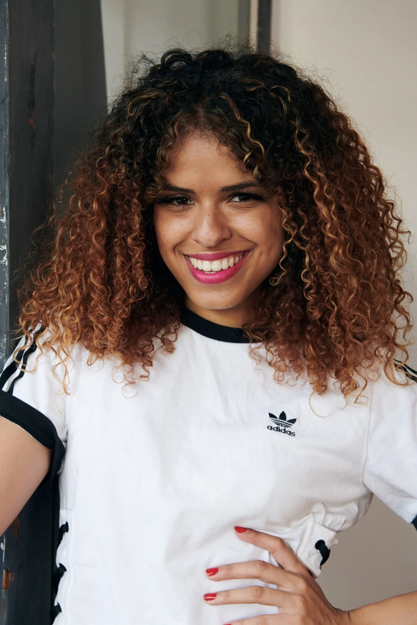 A woman with curly, two-tone hair smiling and wearing a white Adidas t-shirt.