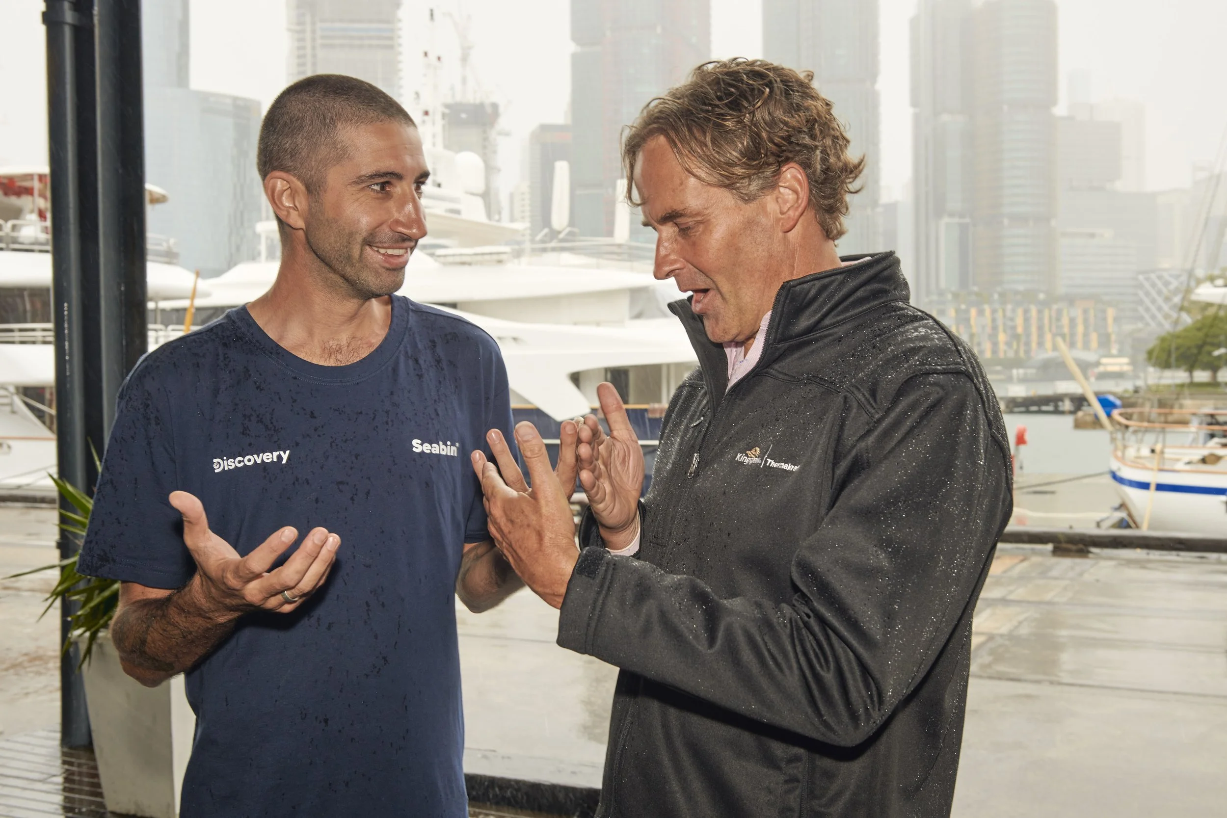 Two men talking outdoors near a marina with yachts and city buildings in the background, one man is wearing a blue shirt data labeled 'Discovery', and the other person is wearing a black jacket with water droplets on it.