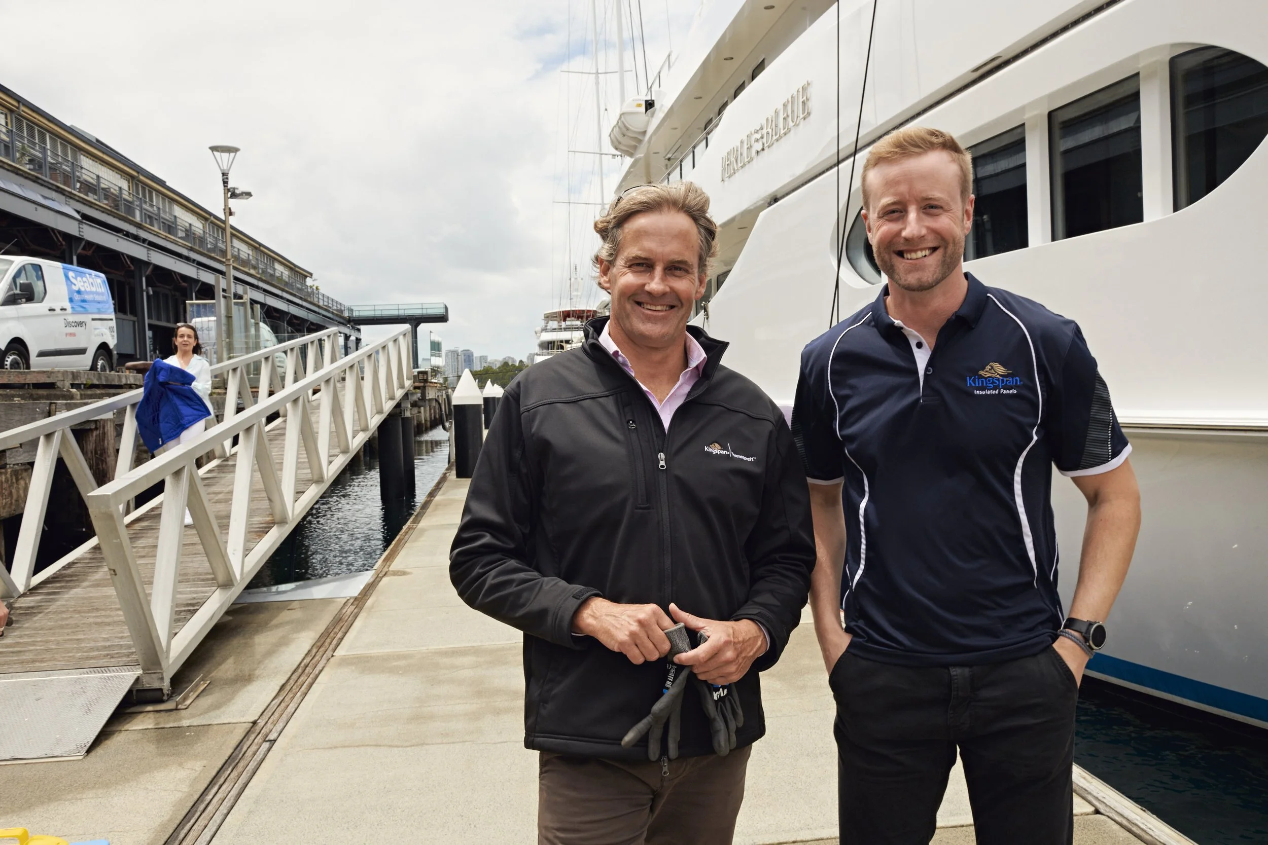 Two men standing on a dock smiling at the camera, with a large yacht behind them and a woman in a blue jacket walking in the background.