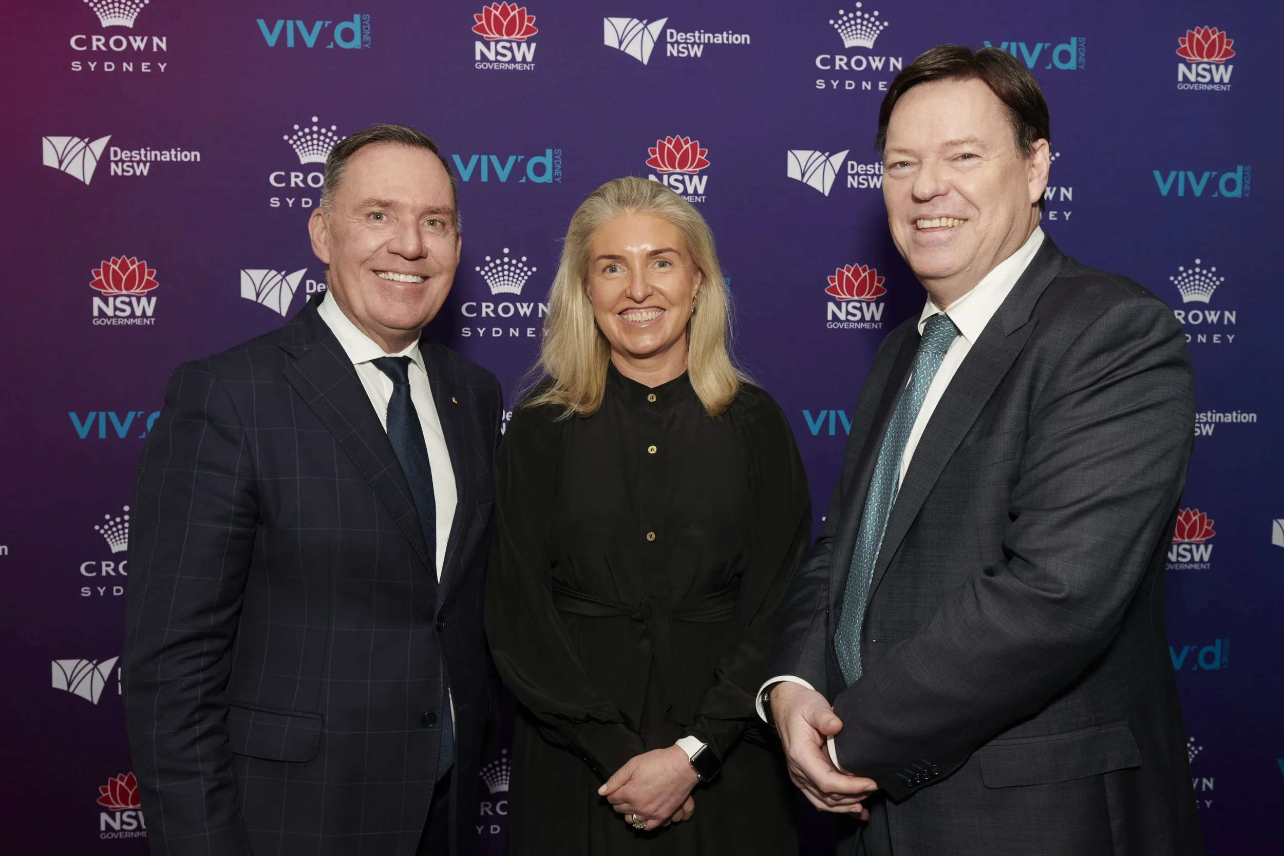 Three professionally dressed people smiling at the camera. Two men in suits and a woman in a black top, standing in front of a purple backdrop with logos including Crown Sydney, VIVID Sydney, and NSW Government.