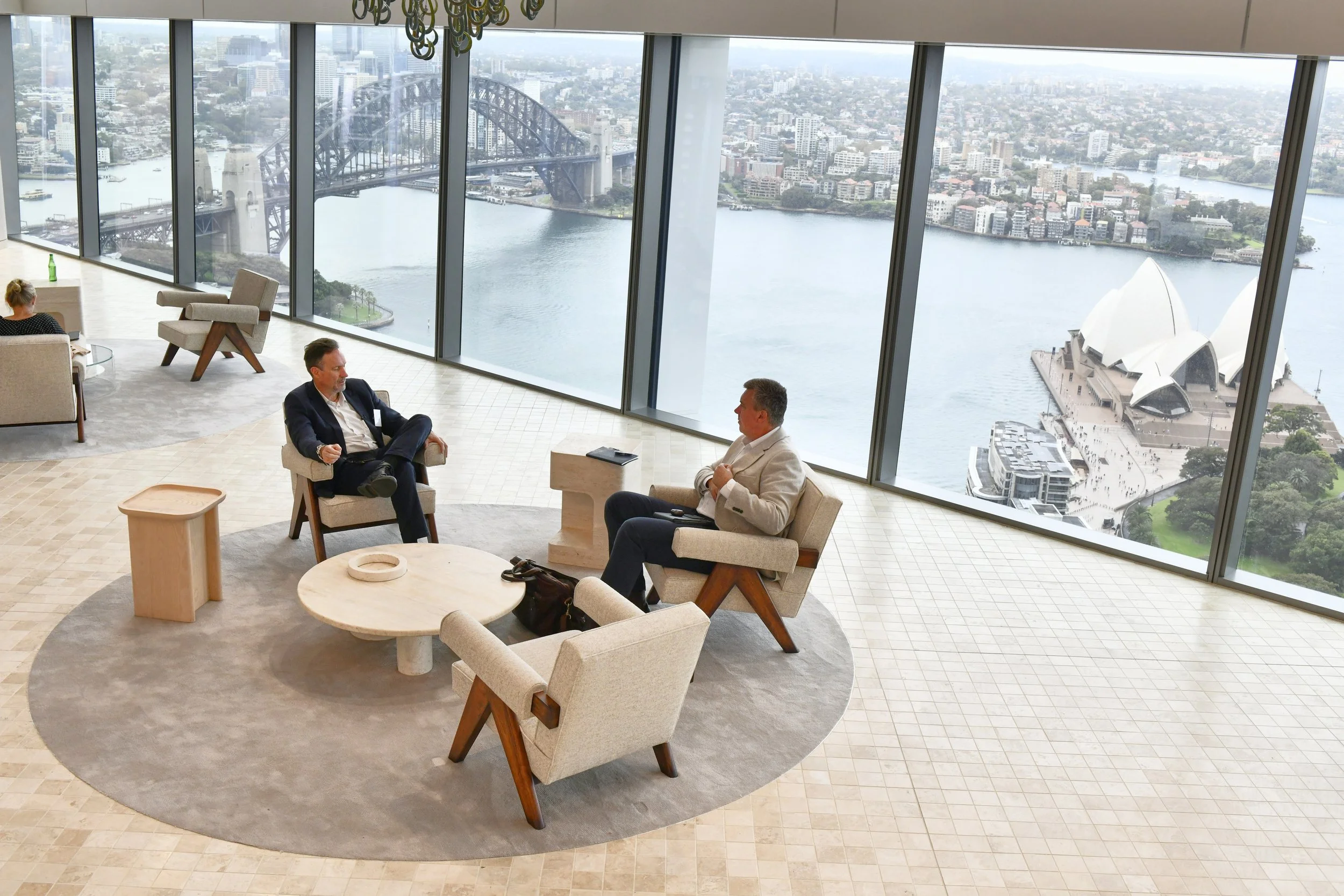Two men sit and converse in an elegant lounge with floor-to-ceiling windows overlooking Sydney Harbour, with the Sydney Opera House and Harbour Bridge visible.