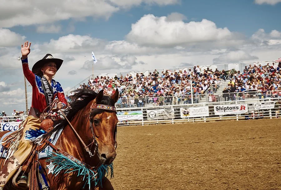 A young woman in a cowboy hat and colorful rodeo outfit riding a brown horse at a rodeo event, waving to the crowd.