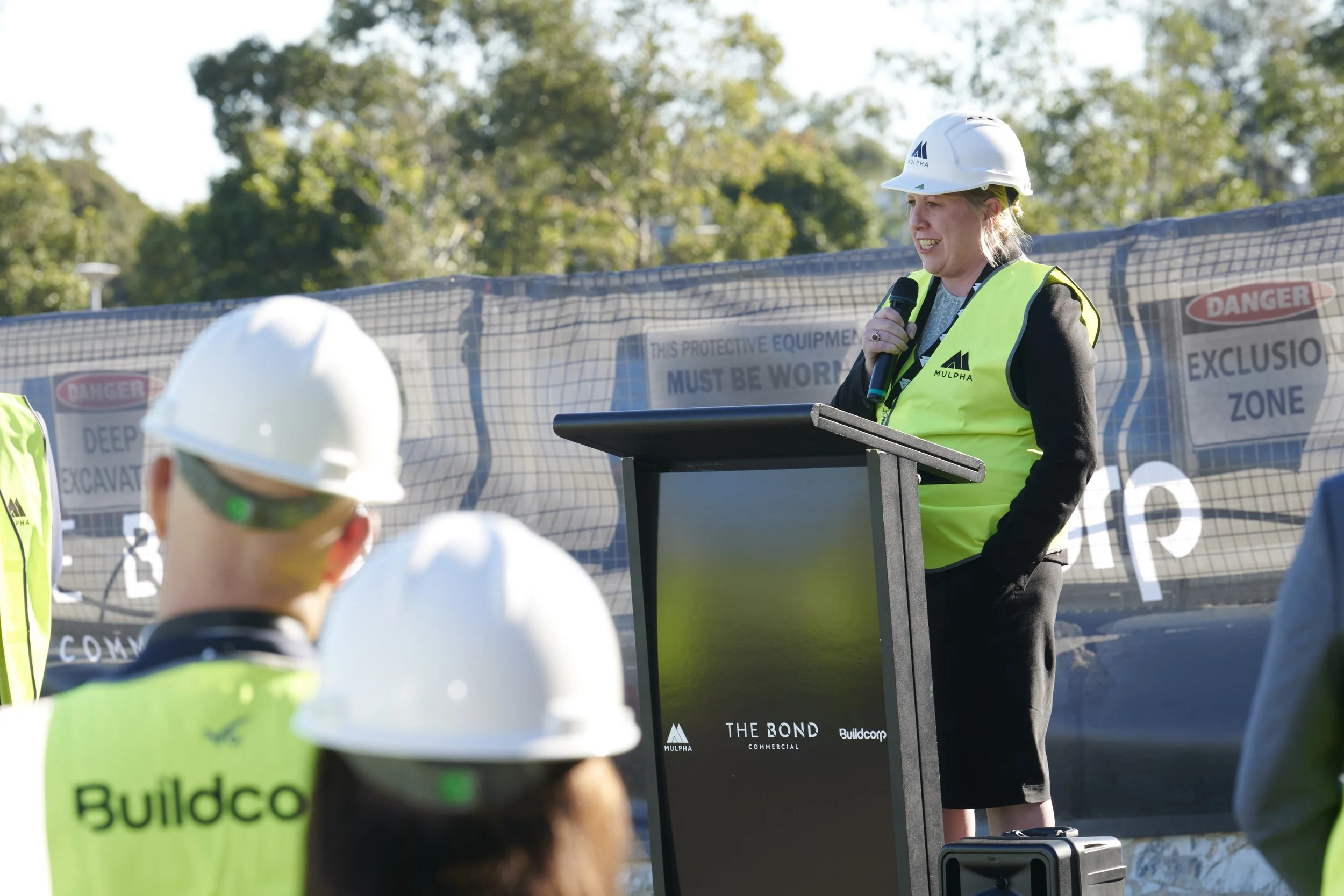 Woman in a safety vest and hard hat speaking at a podium during an outdoor groundbreaking ceremony, with construction workers in helmets listening in the foreground.