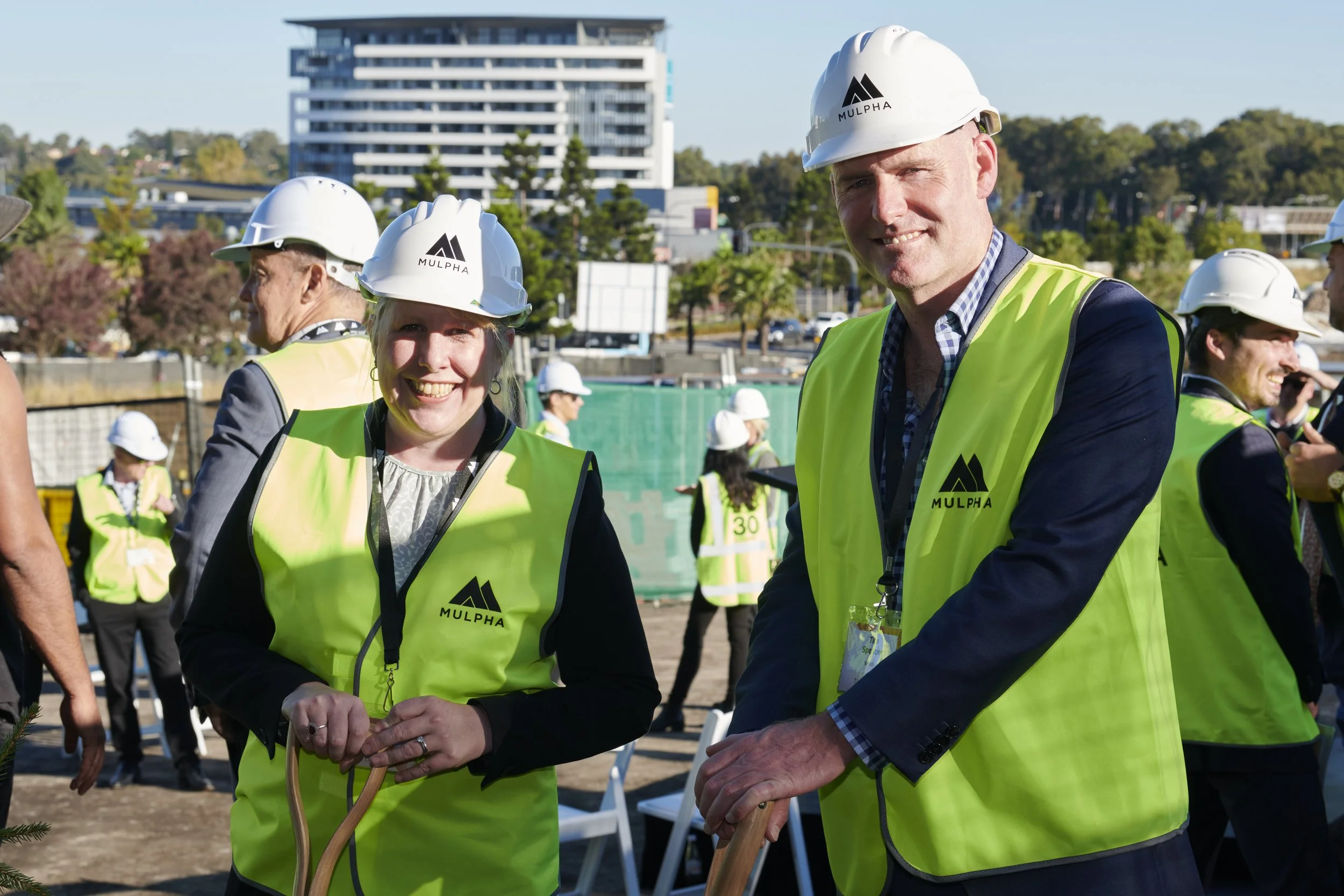 People wearing yellow safety vests and white hard hats at a construction site, some holding shovels. In the background, there are additional workers and a modern building.