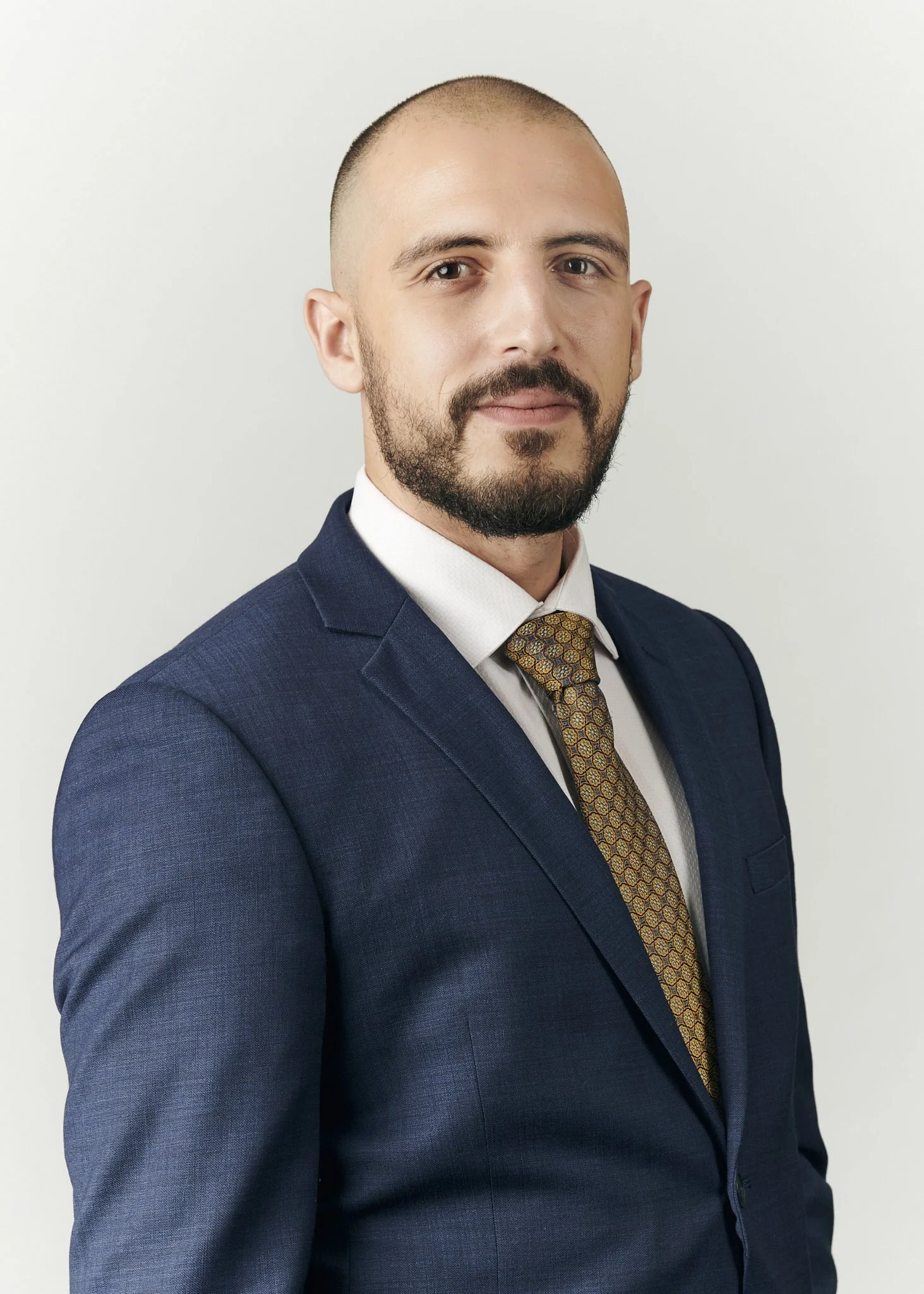 A professional man with a beard and buzz cut hairstyle, wearing a navy blue suit, white dress shirt, and a patterned tie, standing against a plain white background.