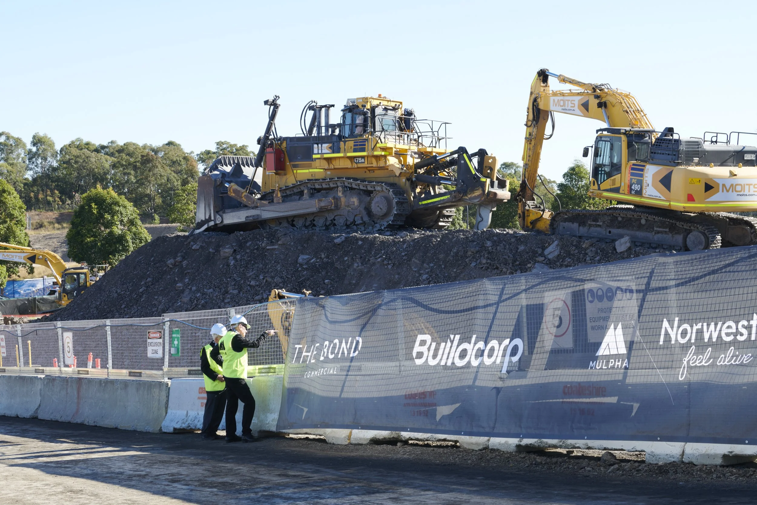 Construction site with heavy machinery, including bulldozer and excavator, on a dirt mound, with a fence and two workers wearing safety helmets and vests in the foreground.