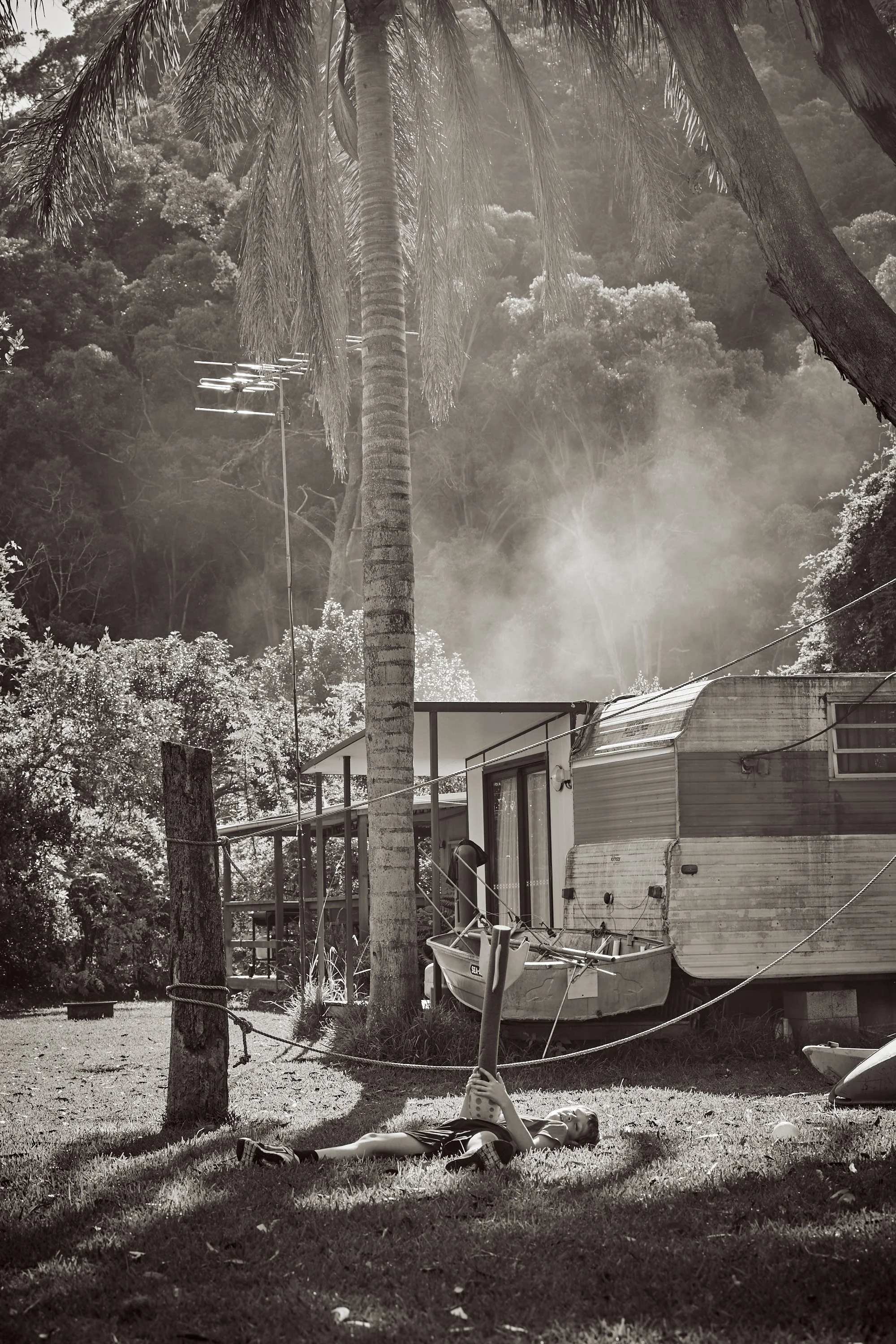 A black and white photo showing a young girl lying on the grass holding a shovel, with a trailer and trees in the background.