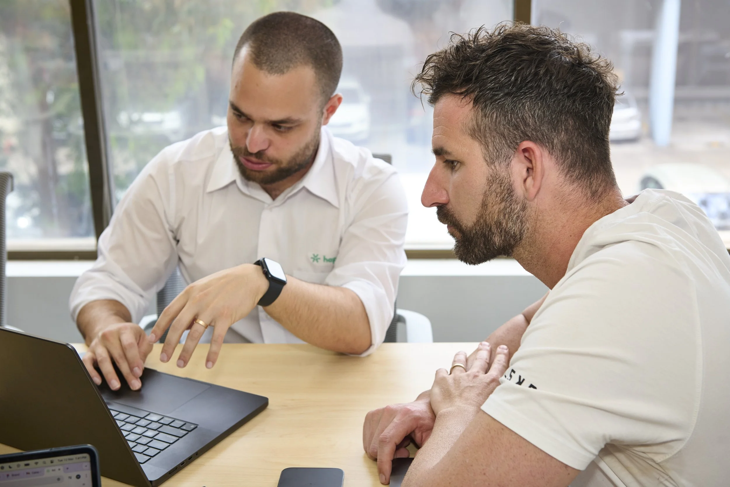 Two men are sitting at a wooden table, engaged in a discussion with a laptop open in front of them. One man is explaining, pointing at the laptop screen, while the other listens attentively, with arms crossed on the table.