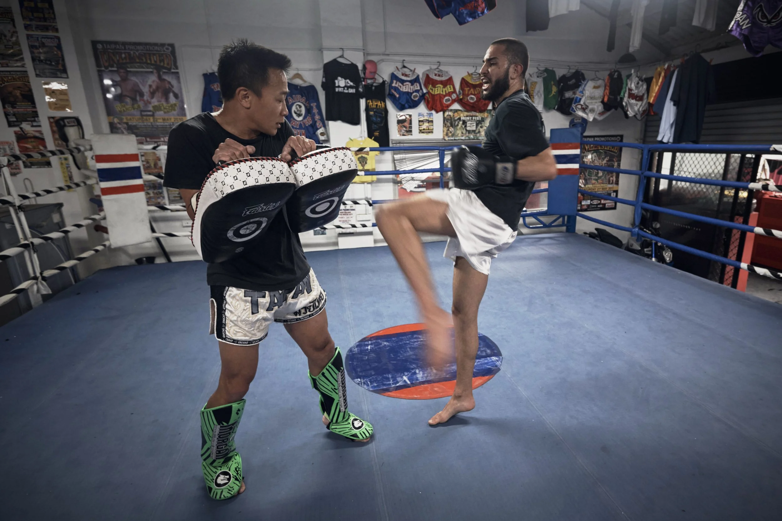 Two men practicing MMA in a boxing gym; one with green MMA gloves and shorts, holding padded targets, and the other executing a knee strike. The gym has various boxing gear and posters on the walls.