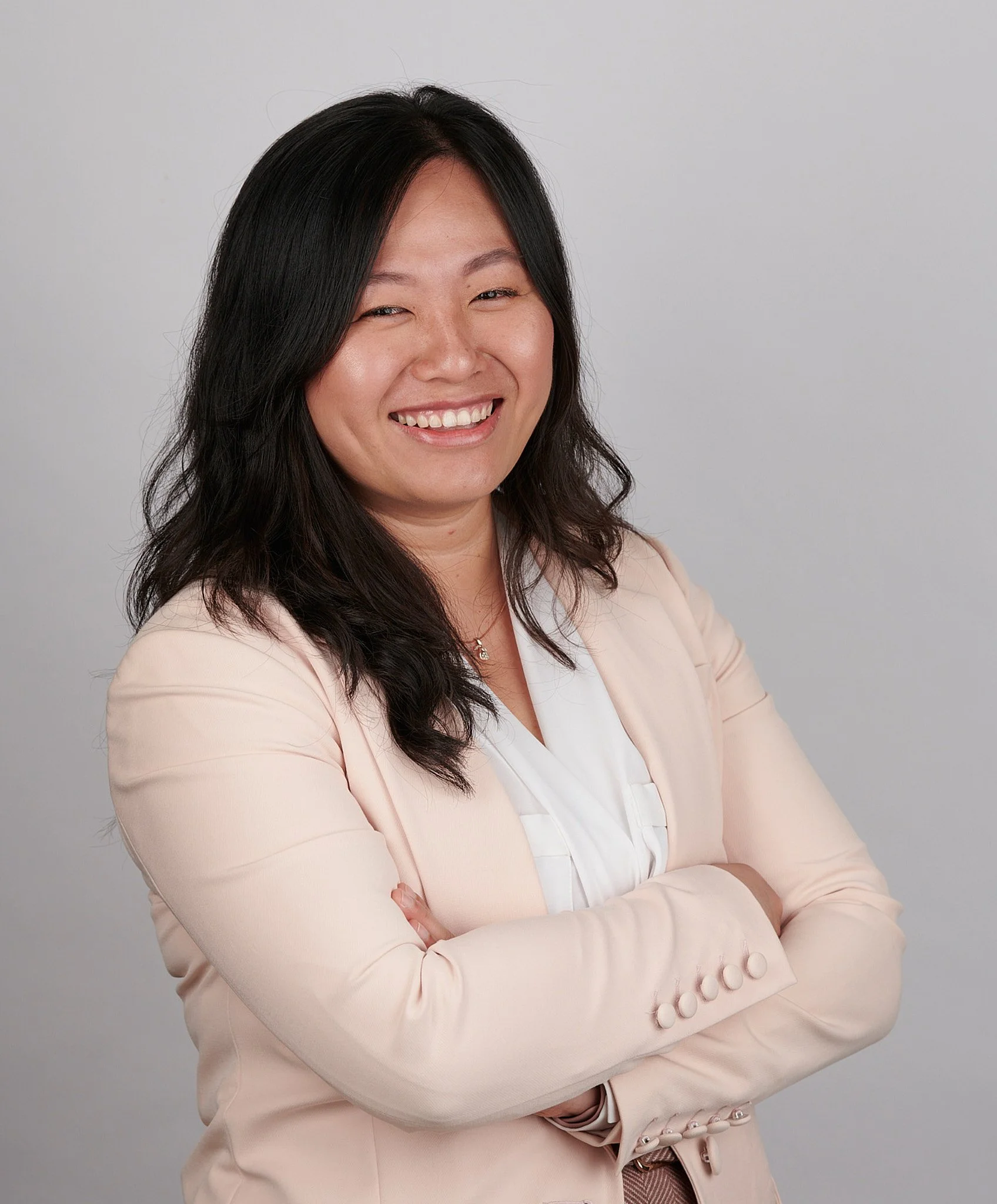 A smiling woman with long dark hair wearing a light-colored blazer and white blouse, standing with arms crossed against a plain gray background.