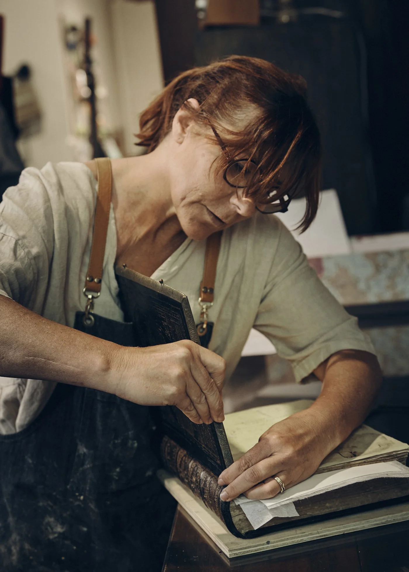 A woman with glasses and a beige shirt, wearing a black apron, is carefully working on an old book in her workshop.