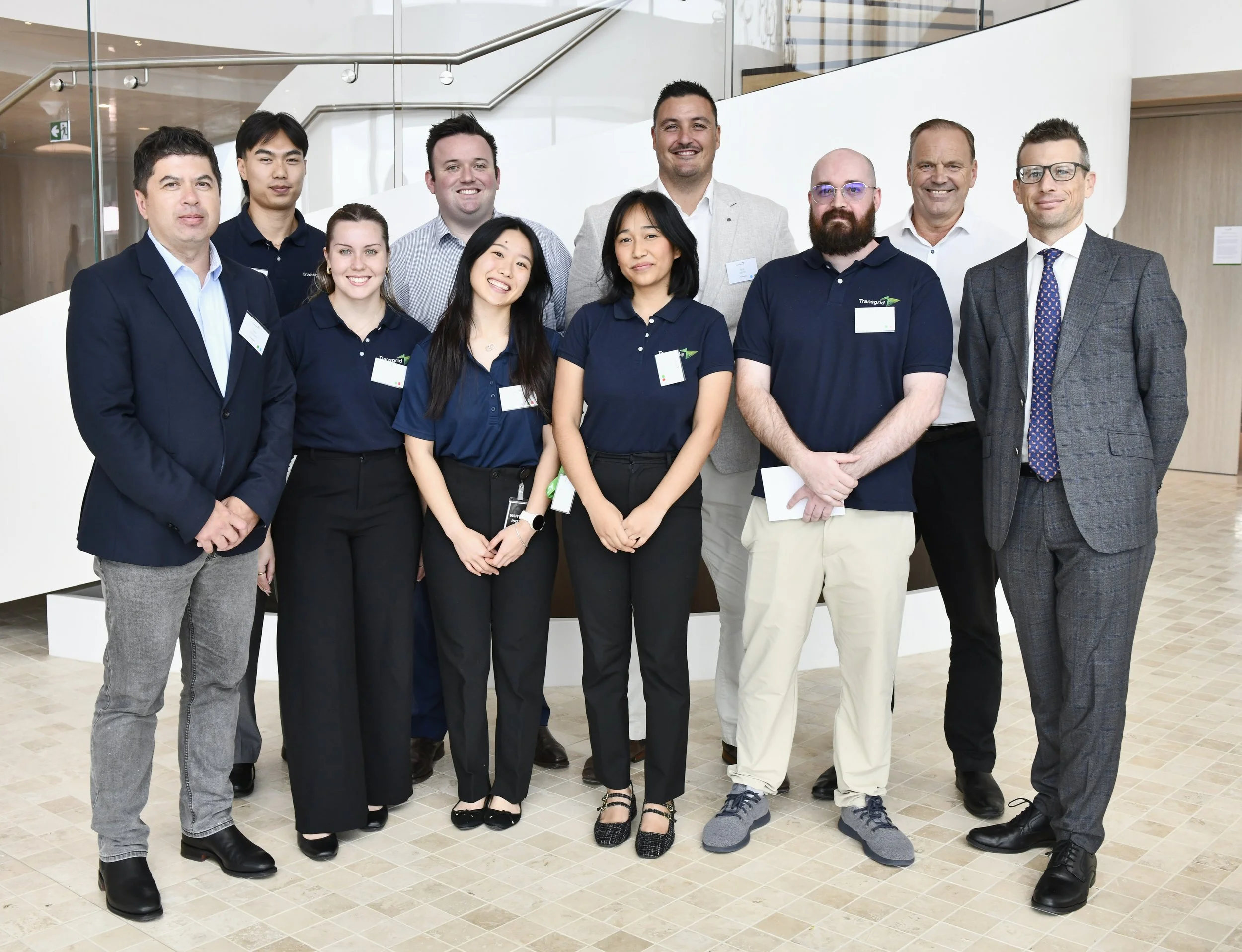 A group of eleven professionals, including six men and five women, posing indoors in an office building for a photo. Some are wearing navy blue polo shirts with a green logo, while others are in business attire. They are smiling and standing in front