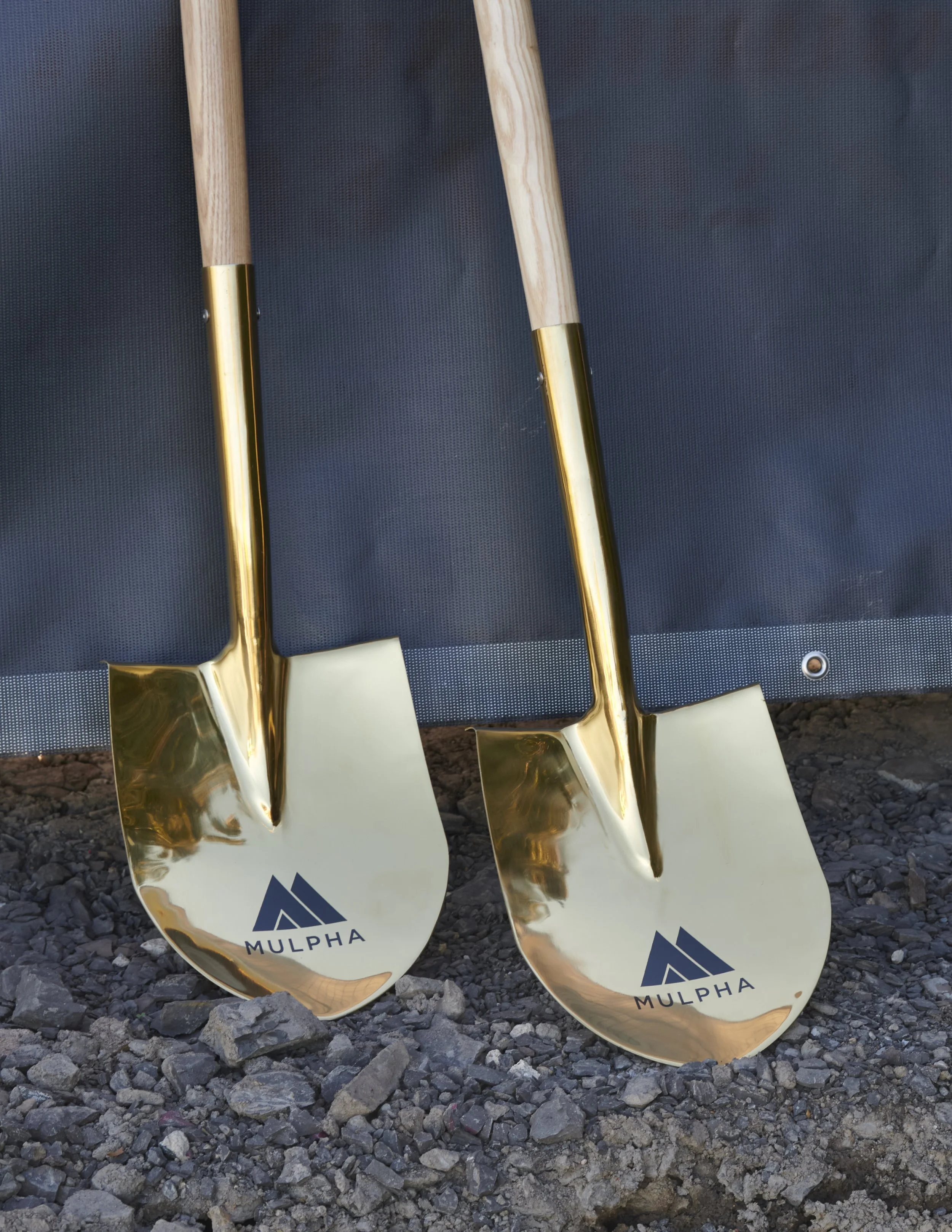 Two gold-colored shovels with wooden handles, positioned side by side on gravel, branded with the MULPHA logo.