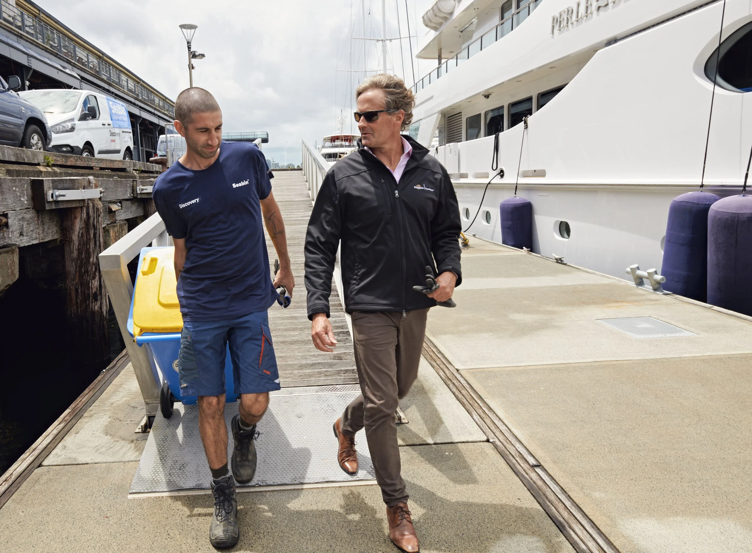 Two men walking on a dock next to a yacht, one in casual clothes and the other in a blue shirt and shorts, carrying a yellow and blue container.