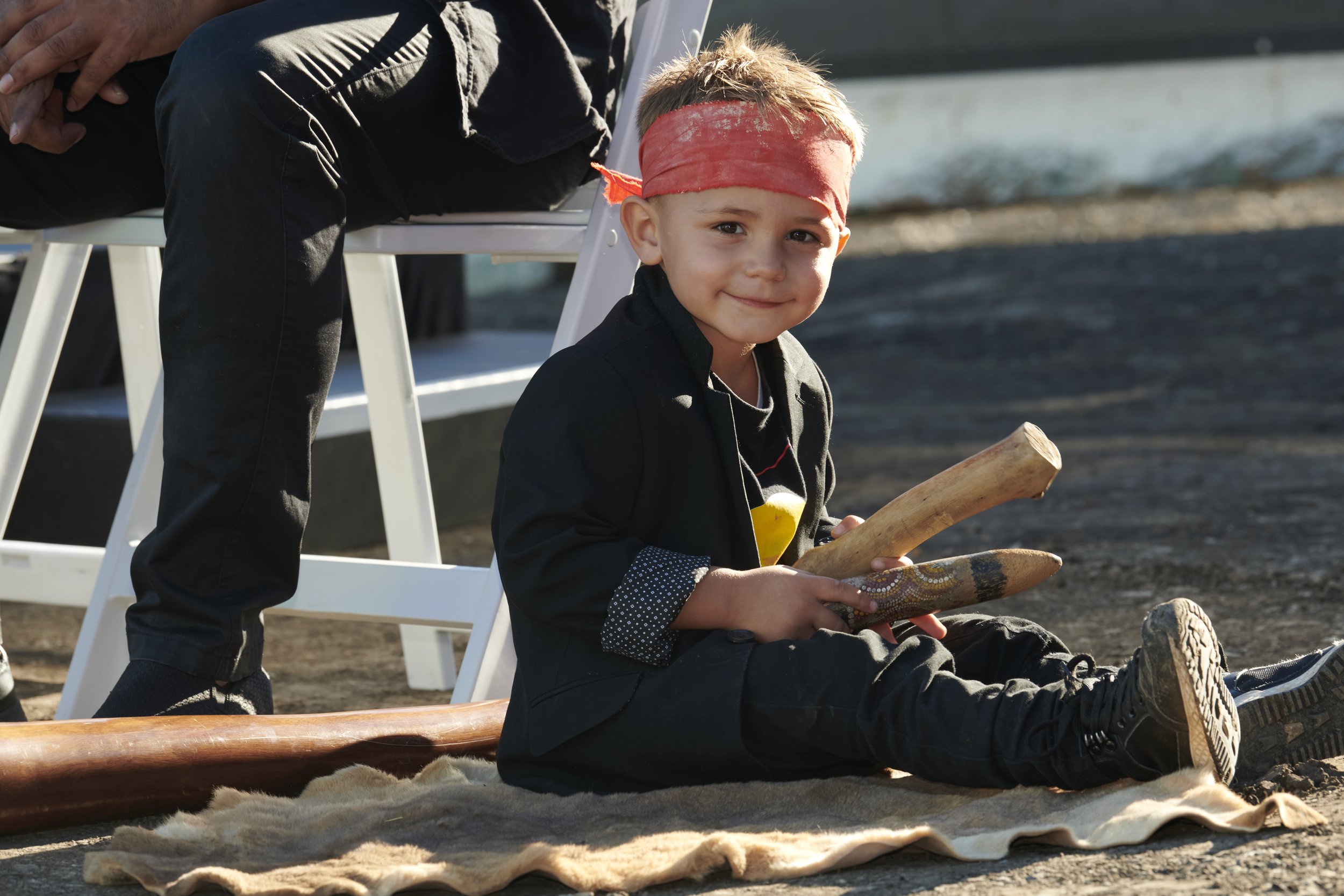 A young boy with a red headband sitting on a blanket outdoors, holding a wooden stick and a decorated wooden object, smiling at the camera.