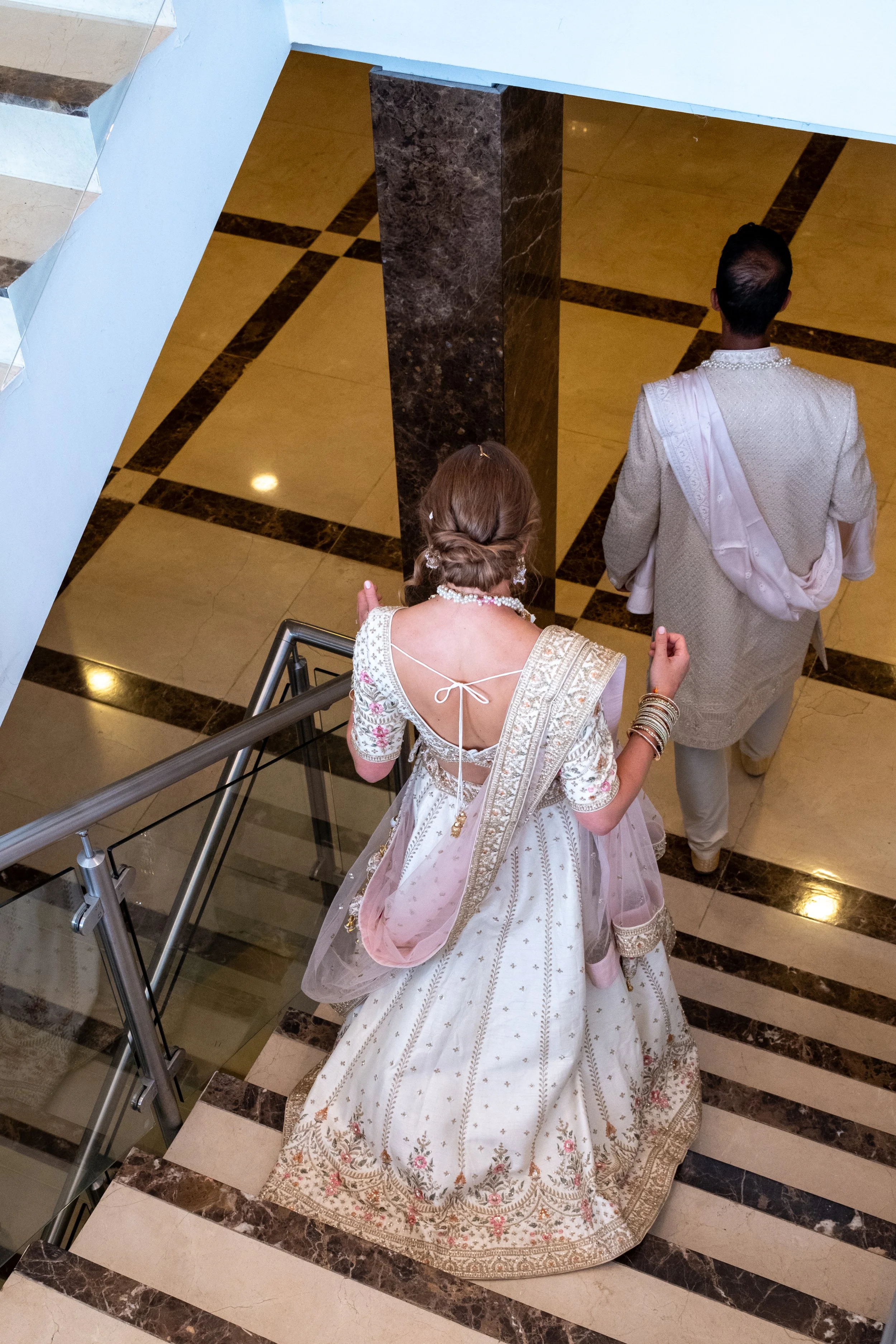 Bride in traditional Indian attire walking down a staircase behind the groom in a cream-colored sherwani en route to the reception