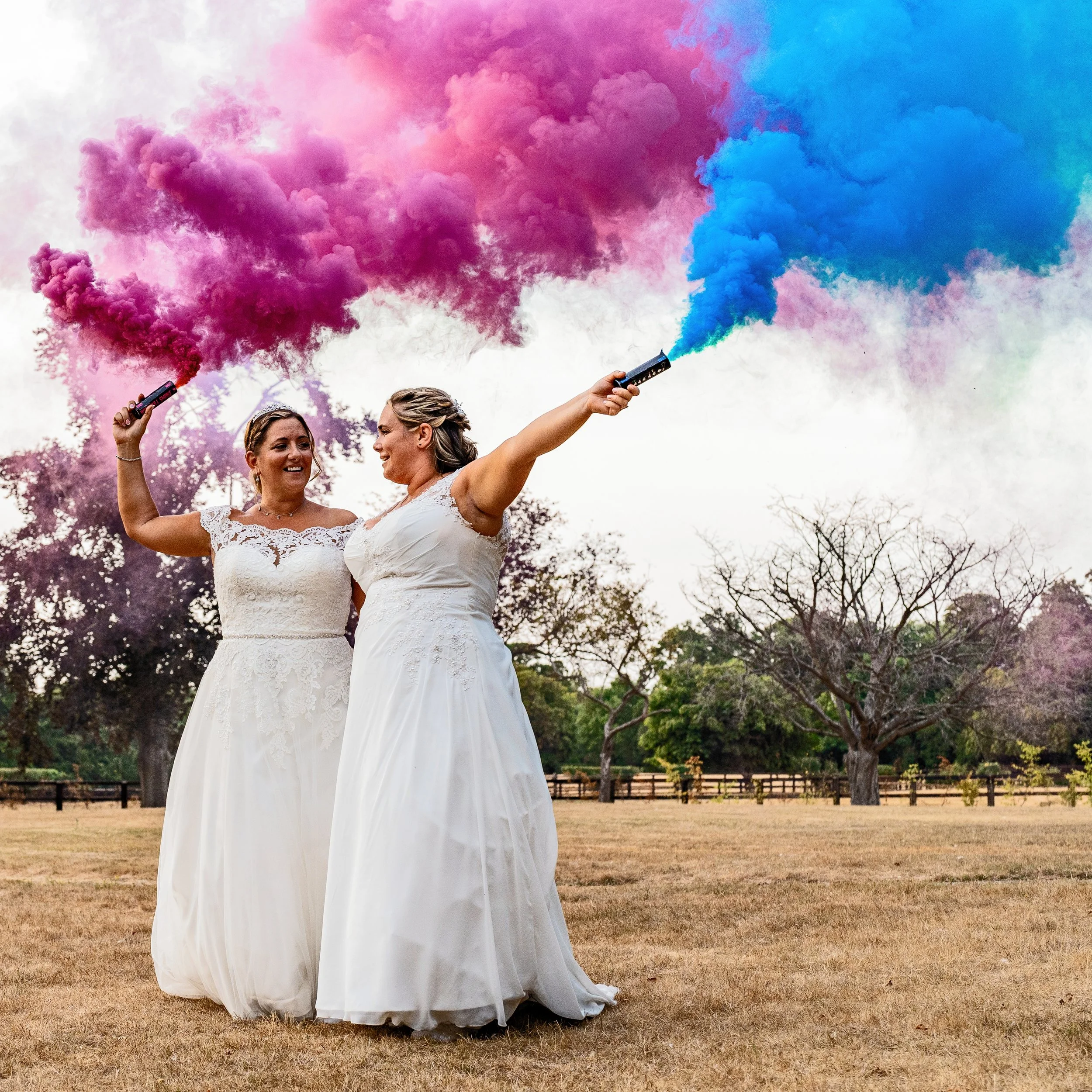 Two women in wedding dresses holding smoke bombs that emit pink and blue smoke during an outdoor celebration 