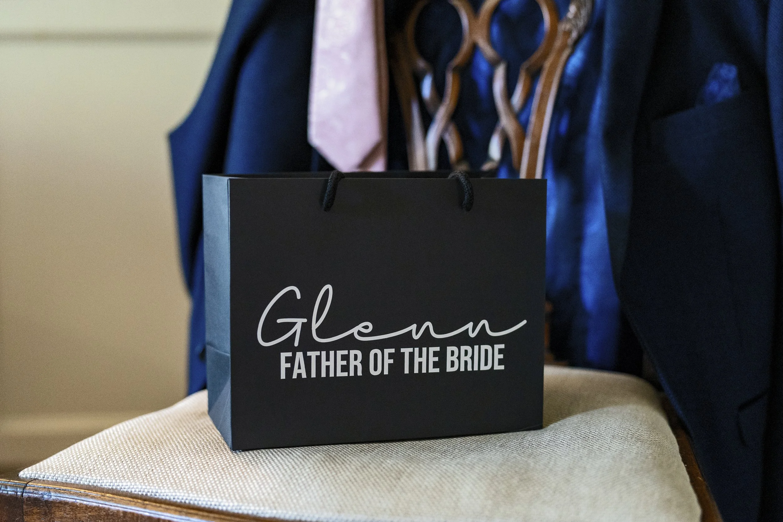 A black gift bag with white text that reads 'Groom, Father of the Bride' sitting on a padded beige surface, with a person in a dark suit and pink tie standing behind it. The focus is on the gift bag.