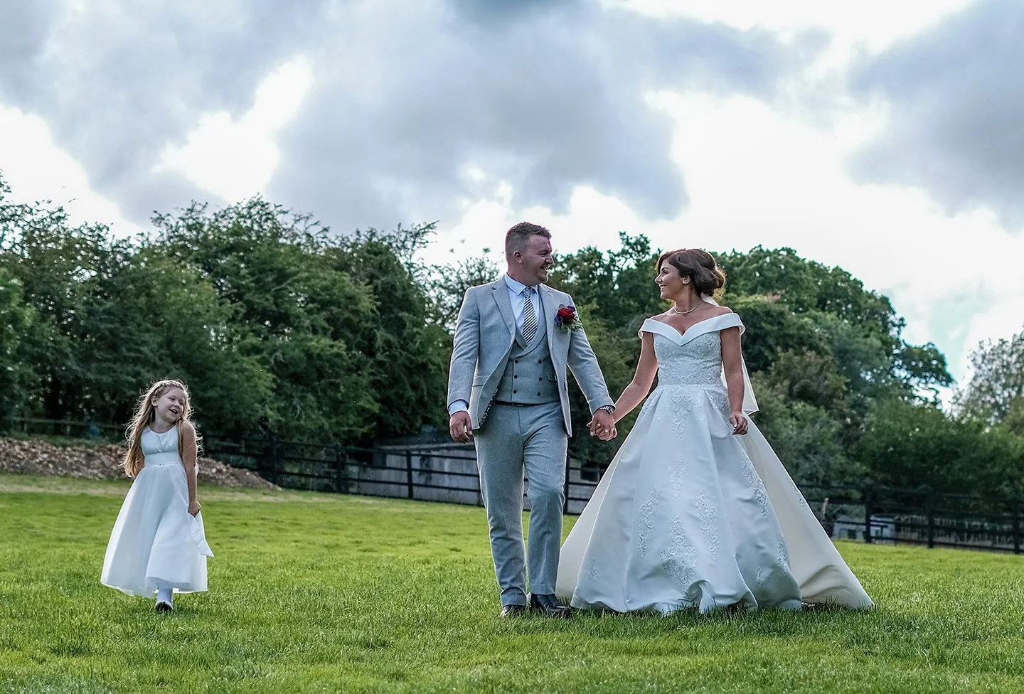 A bride and groom holding hands, walking on grass outdoors with a young girl, all dressed in wedding attire, against a backdrop of trees and a cloudy sky.