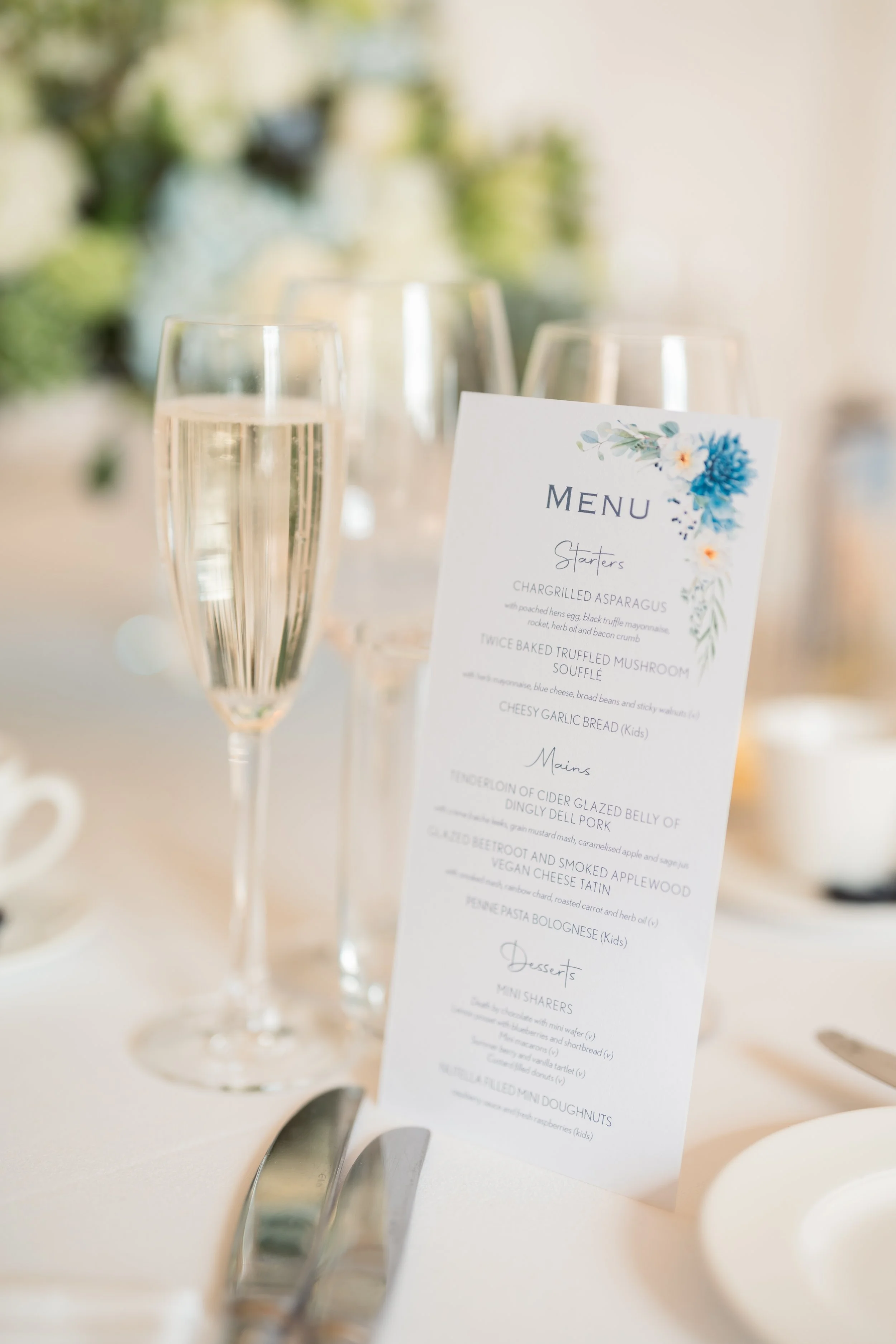 A wedding menu card standing on a table with champagne glasses and utensils, set in a well-lit environment with floral decorations.