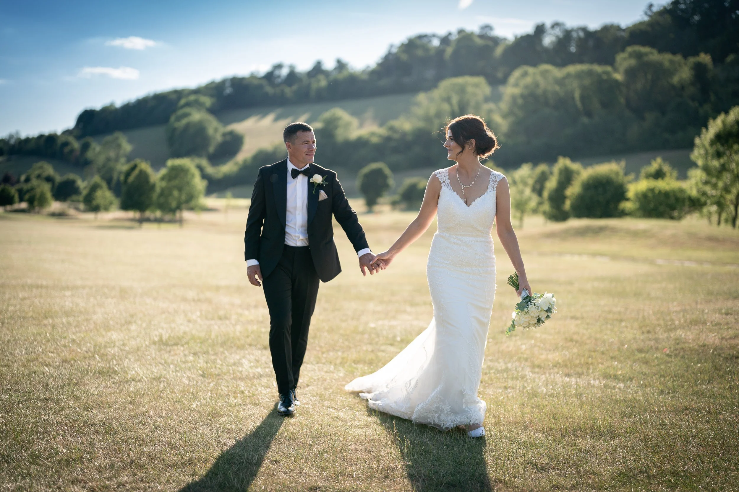 A newlywed couple walking hand in hand on a grassy field 