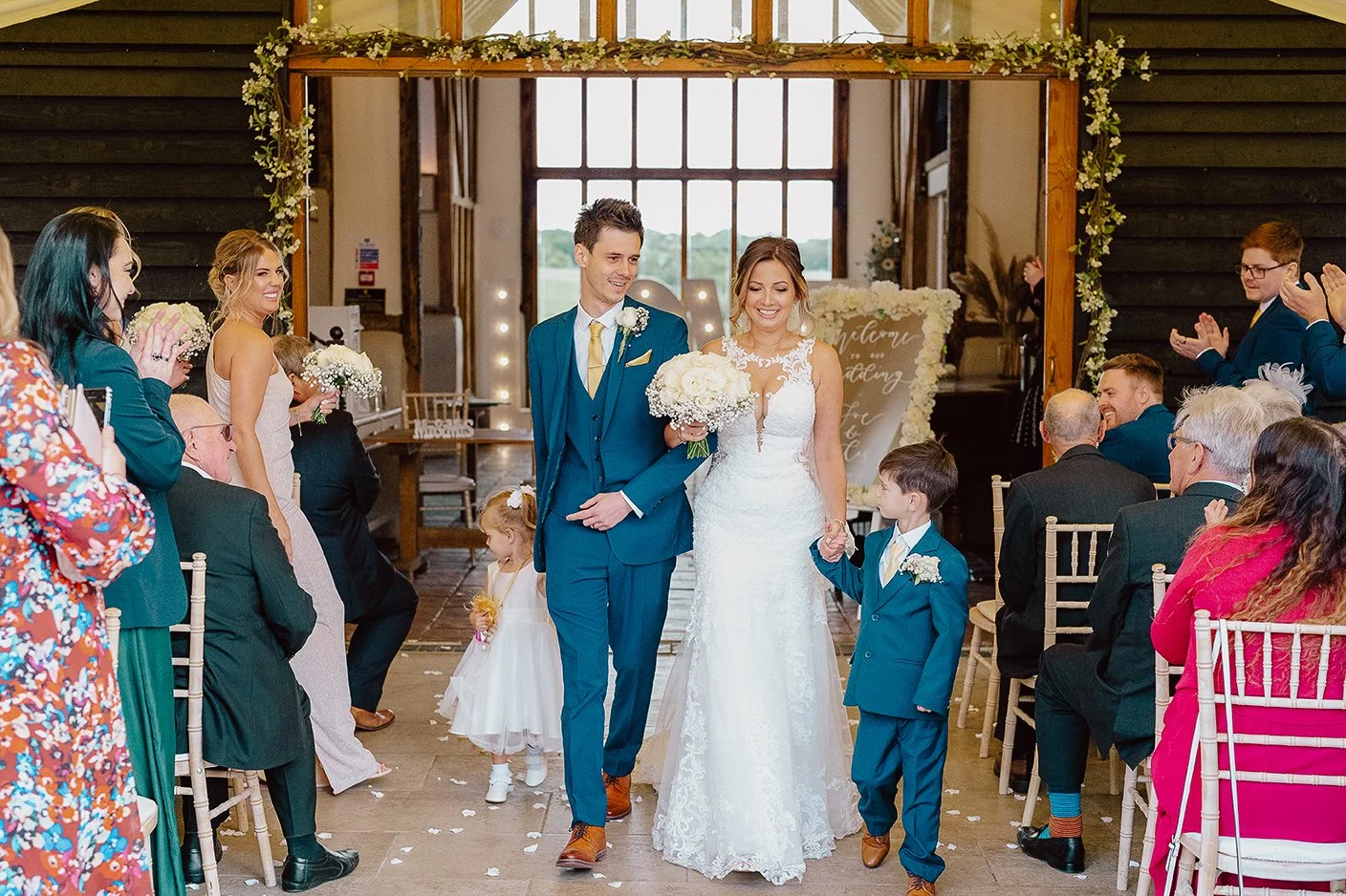 A bride and groom walking down the aisle with two young boys holding their hands, surrounded by clapping guests at a wedding ceremony in a rustic venue with large window and floral decor.