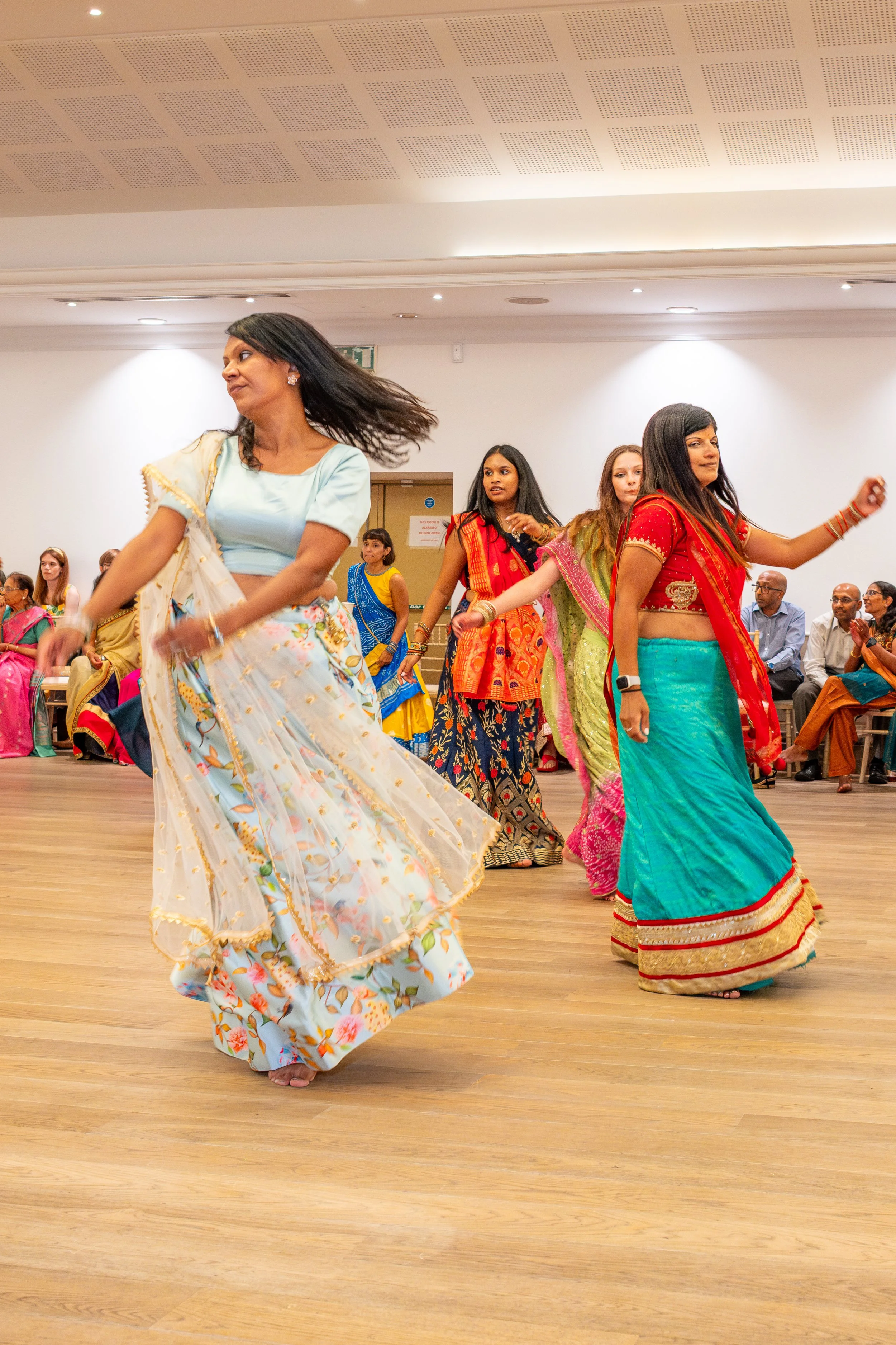 Women dancing in traditional Indian sarees in a hall with seated audience at an Indian wedding