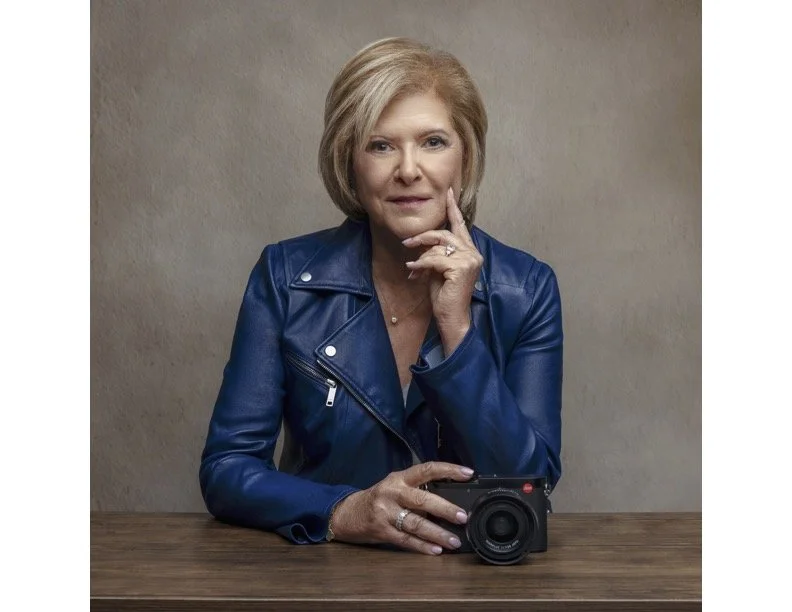 A color photograph of the photographer, Lisa A. Cutler, looking straight at the camera, wearing a blue leather jacket, and resting her hand on a Leica camera.