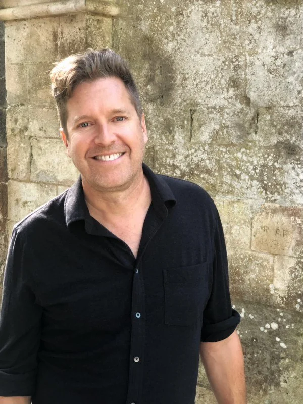 A smiling man with short brown hair, wearing a black shirt, standing outdoors in front of a weathered stone wall.