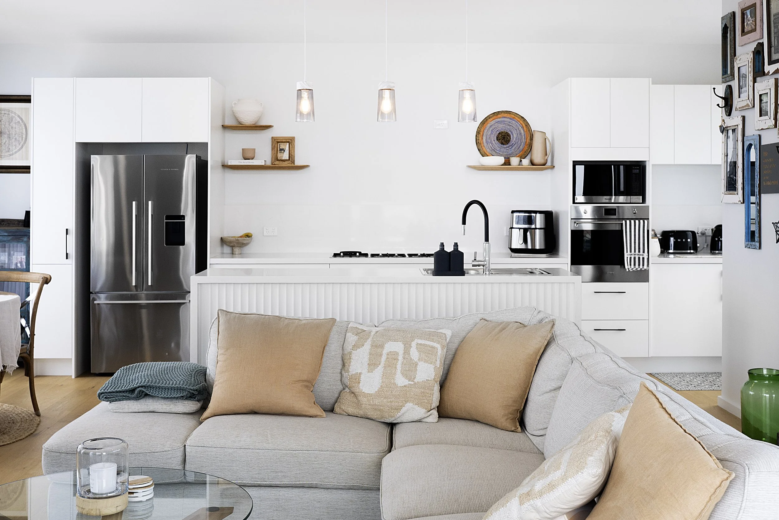 A modern kitchen with white cabinets, stainless steel refrigerator, open shelving, pendant lighting, and a living area with a beige sofa, pillows, and a glass coffee table in cronulla. 