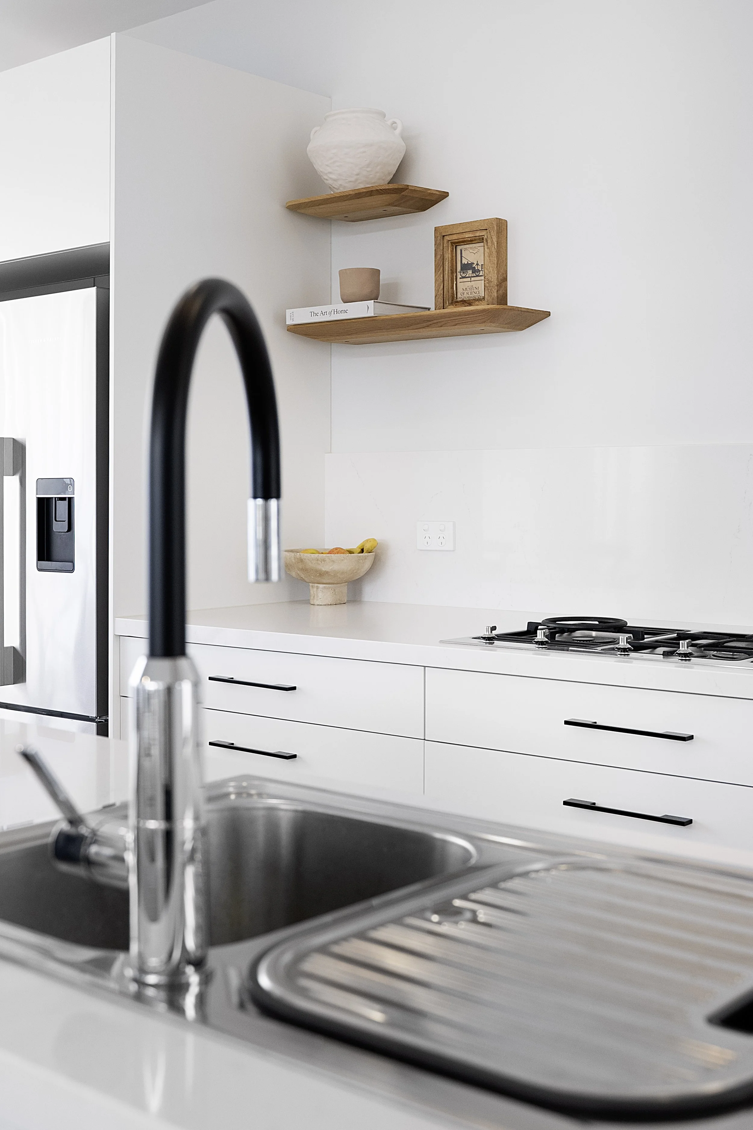 Modern kitchen with white cabinetry, a stainless steel sink, a black faucet, again stainless steel stove, wooden floating shelves with decorative items, a bowl of fruit, and a white wall with electrical outlets in cronulla. 