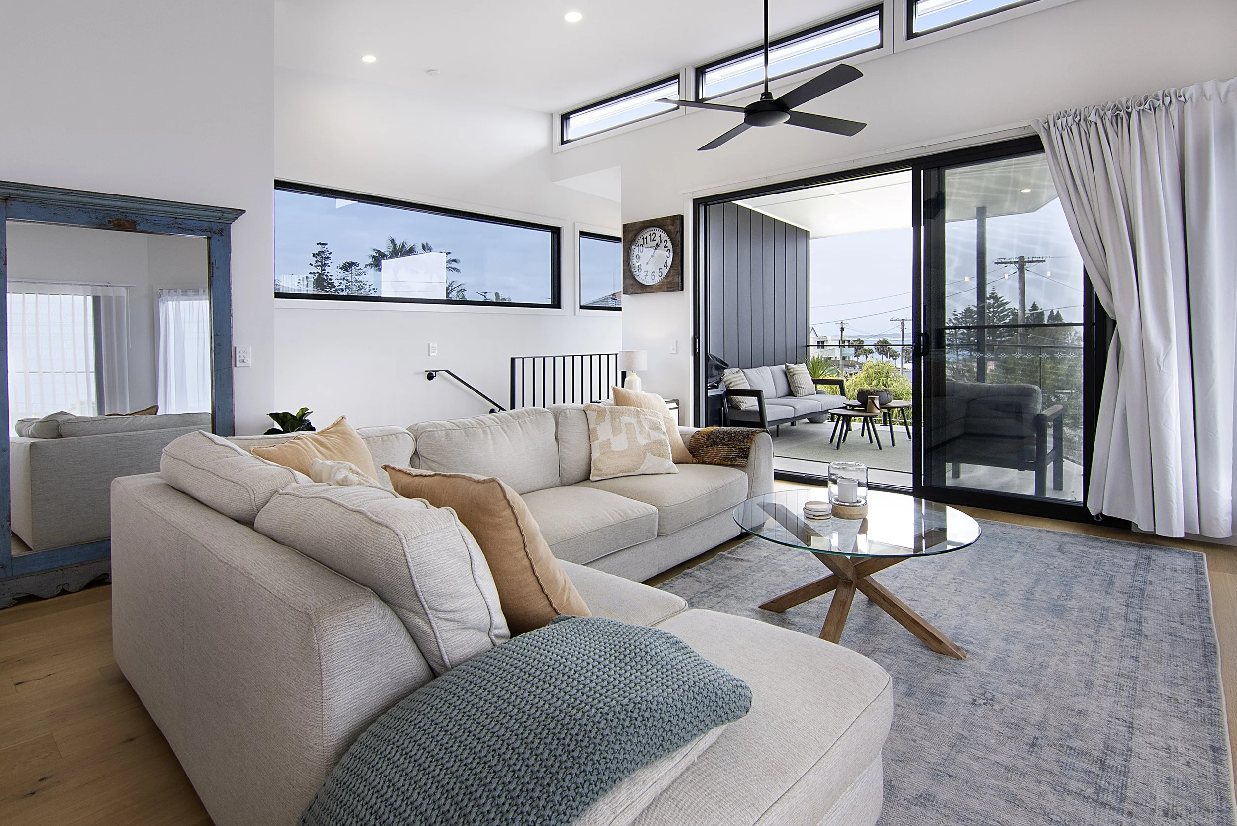 Modern living room with white sofa, beige cushions, glass coffee table, patterned rug, large windows revealing outdoor scenery, sliding glass door, ceiling fan, wall clock, and balcony area in cronulla. 