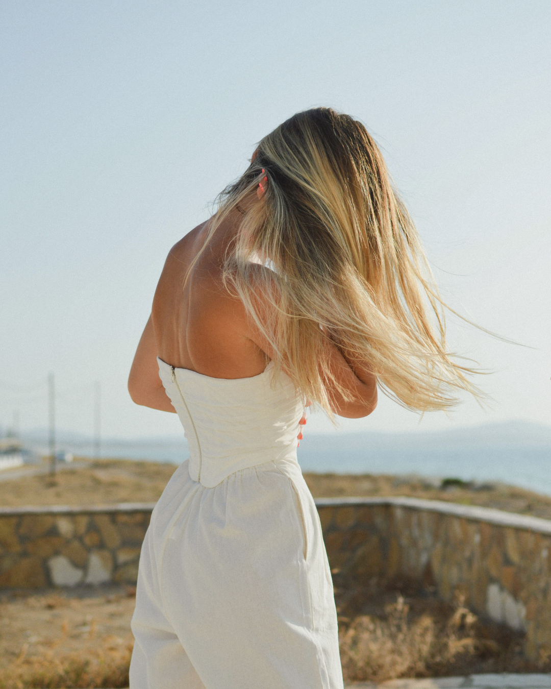 Photograph of a woman standing outdoors near the coast, viewed from behind, with one hand resting near her neck and shoulder as her hair moves in the wind, suggesting subtle tension through the neck and upper back.