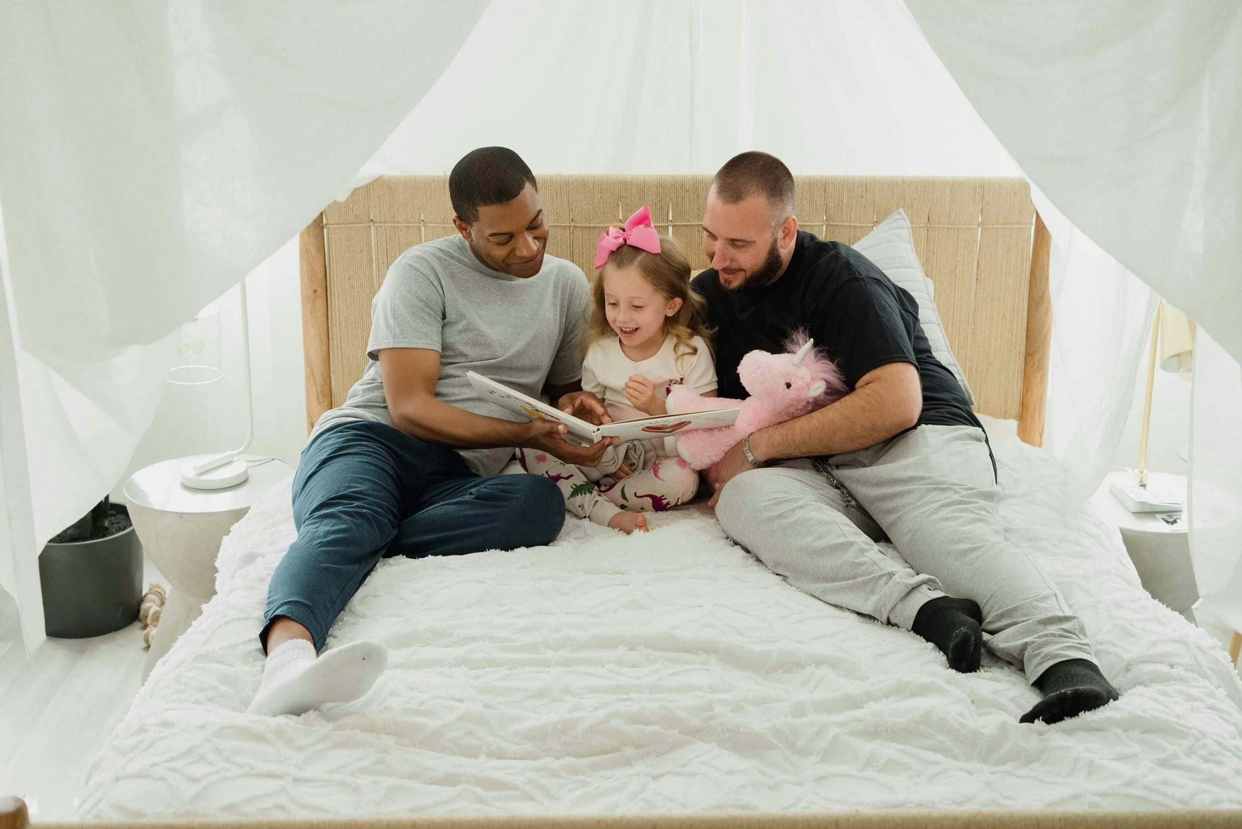 Two fathers reading a book with their young daughter on a bed, representing LGBTQ family planning and fertility support at South Shore Acupuncture and Wellness, Rockland MA.