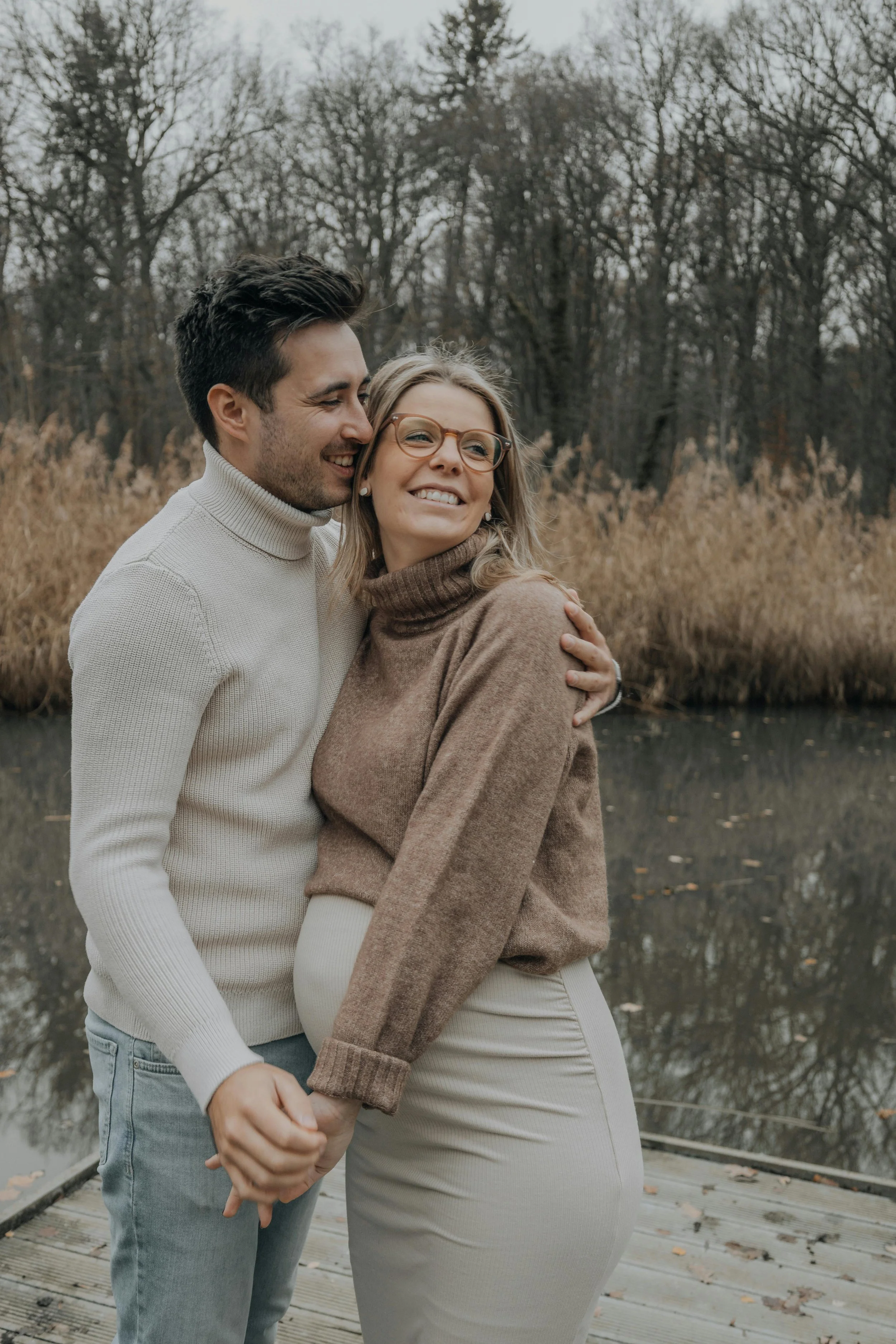 A pregnant woman and her partner laughing together on a waterfront dock in autumn, representing fertility and IVF support at South Shore Acupuncture and Wellness, Rockland MA.