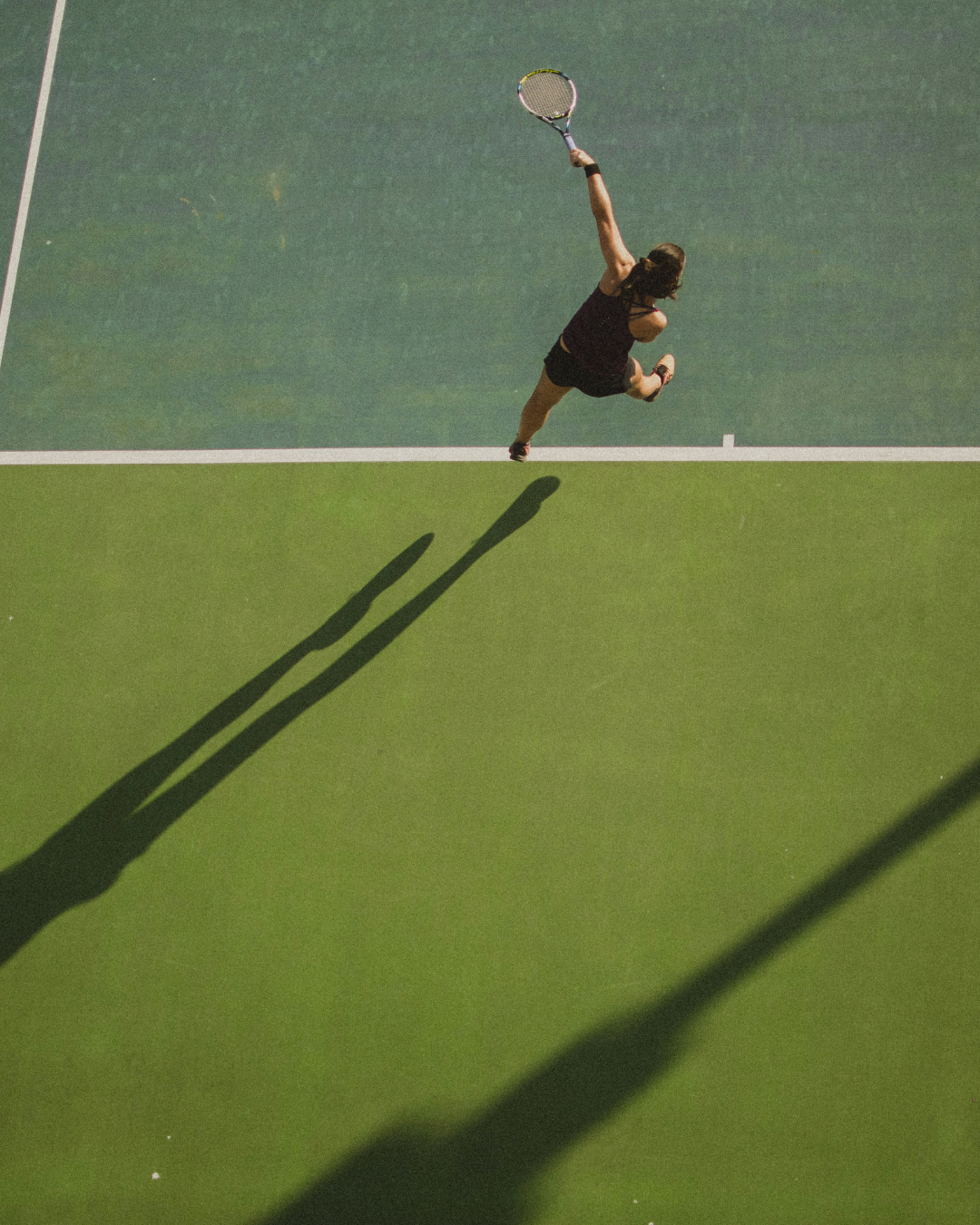 Overhead photograph of a person serving a tennis ball on an outdoor court, arm extended upward with a racket, emphasizing repetitive use of the elbow, wrist, and hand during forceful movement.