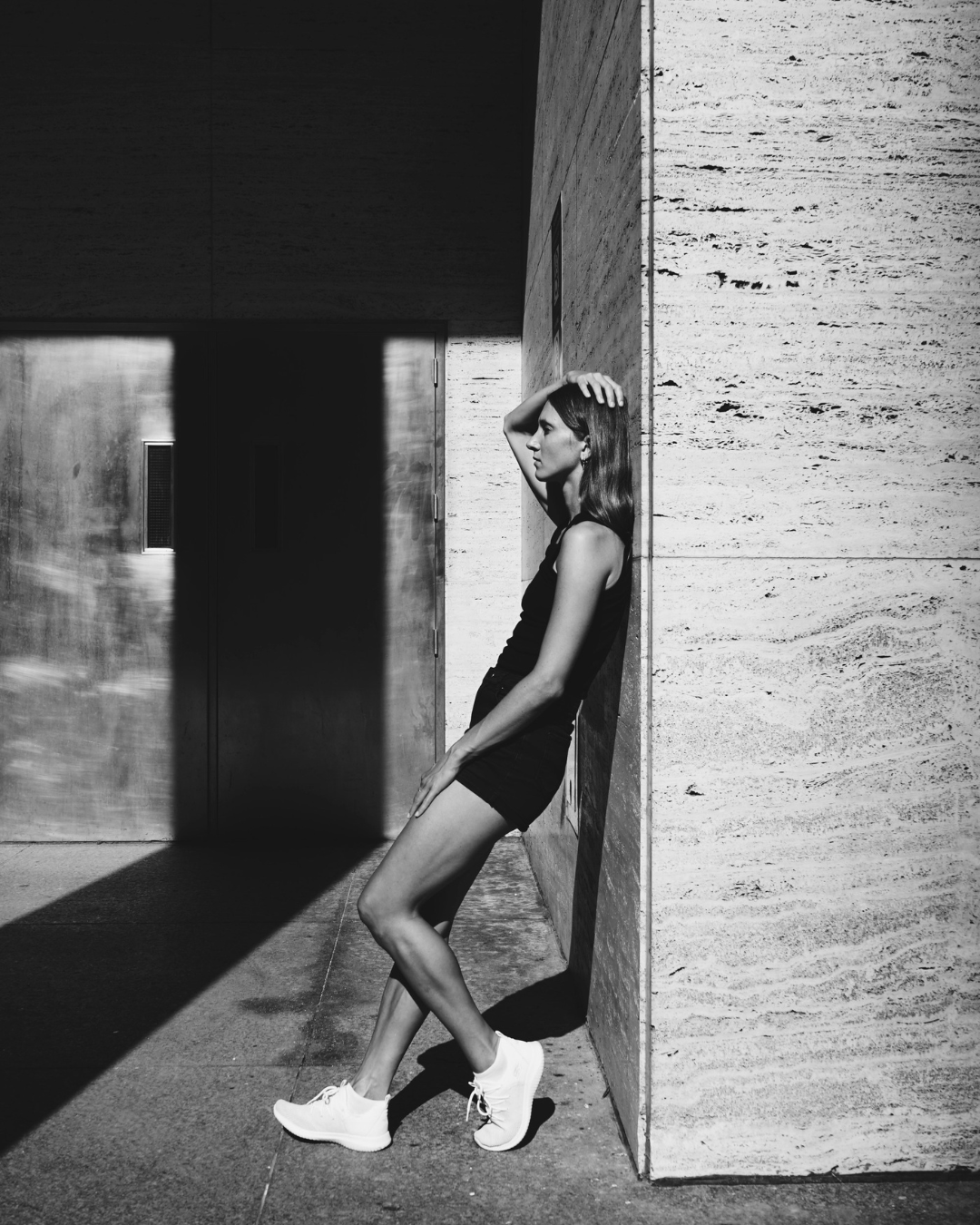 lack and white photograph of a woman leaning back against a stone wall in an urban setting, one arm resting on her head, her body partially in shadow, suggesting physical strain and quiet tension.