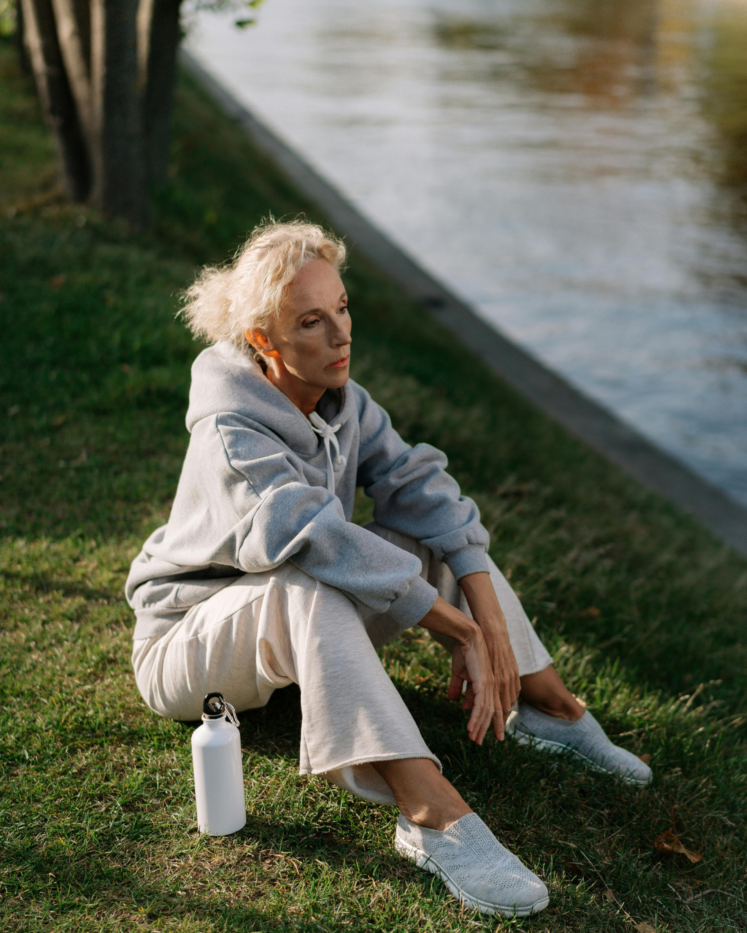 Photograph of an older woman sitting on grass near a body of water, knees bent and arms resting on her legs, suggesting quiet rest and awareness of knee discomfort during everyday movement.