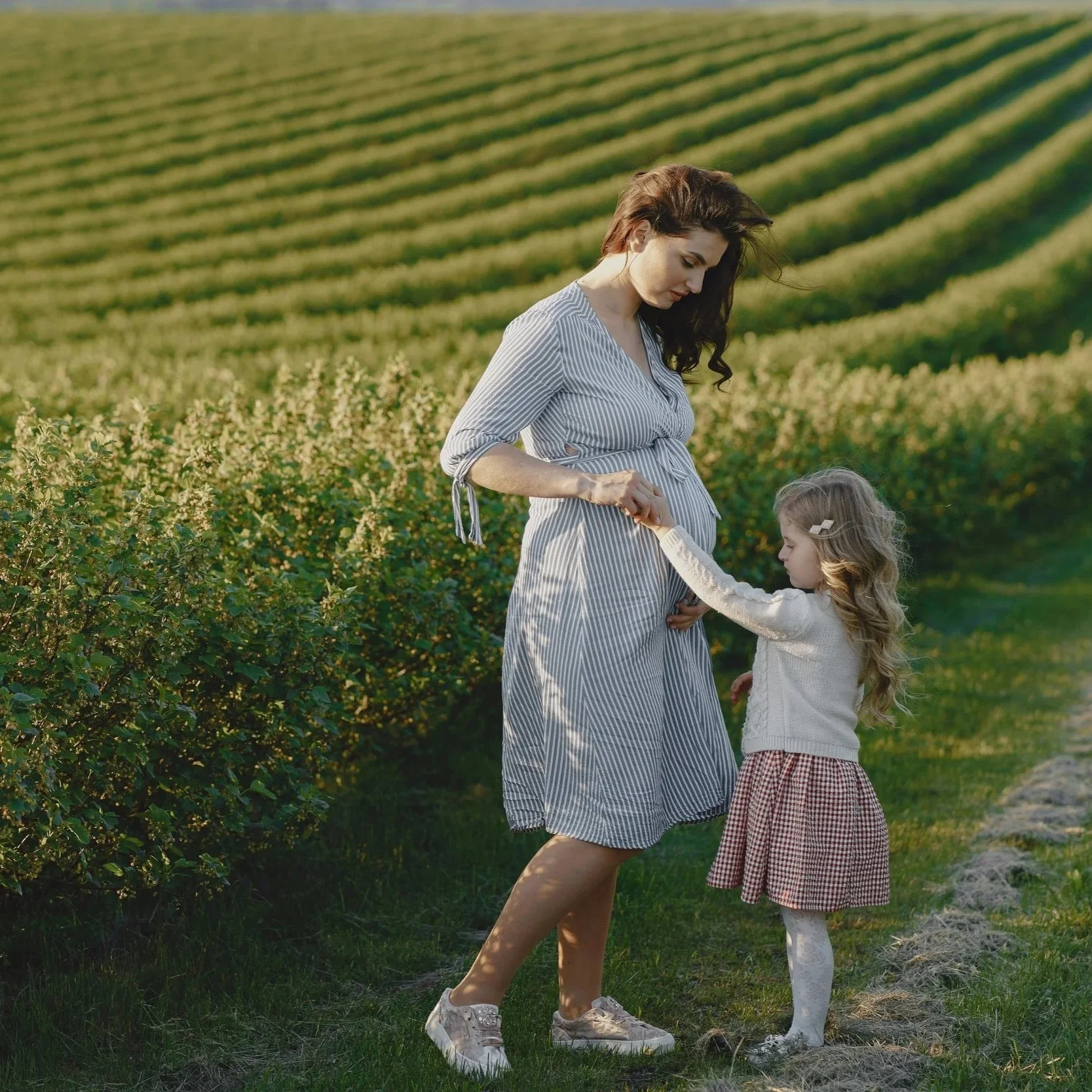 Pregnant woman in a striped dress holding her young daughter's hand along a sunlit garden path, representing prenatal care and fertility support at South Shore Acupuncture and Wellness, Rockland MA.