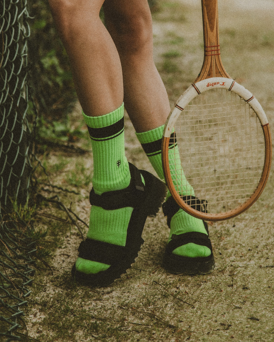 Close up photograph of a person’s lower legs and feet standing on a sandy surface, wearing sandals over socks and holding a tennis racket, emphasizing the feet bearing weight and absorbing impact during repeated use.