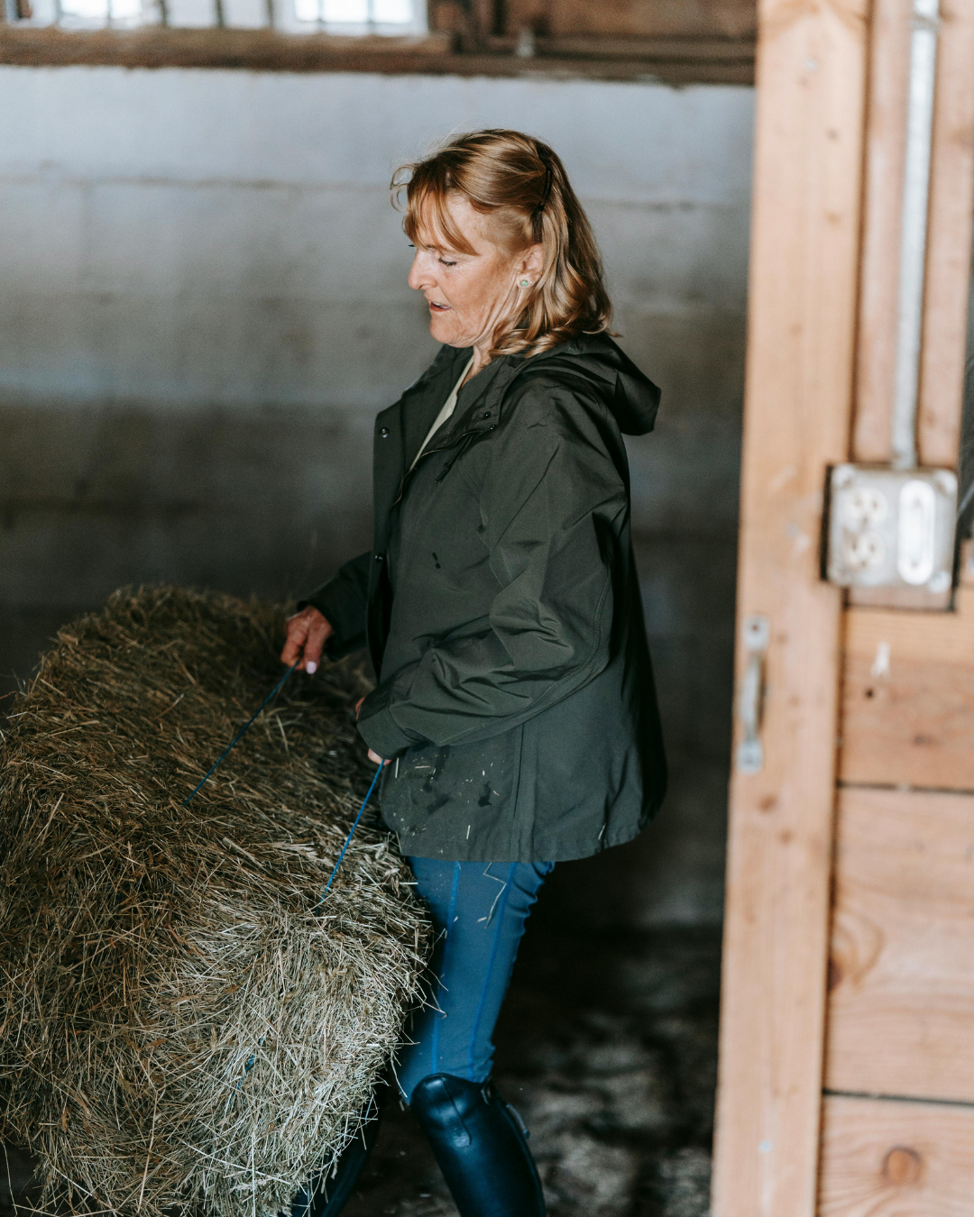 Photograph of a woman standing indoors beside a bale of hay, slightly bent forward while working, emphasizing load through the lower back and hips during everyday physical activity.