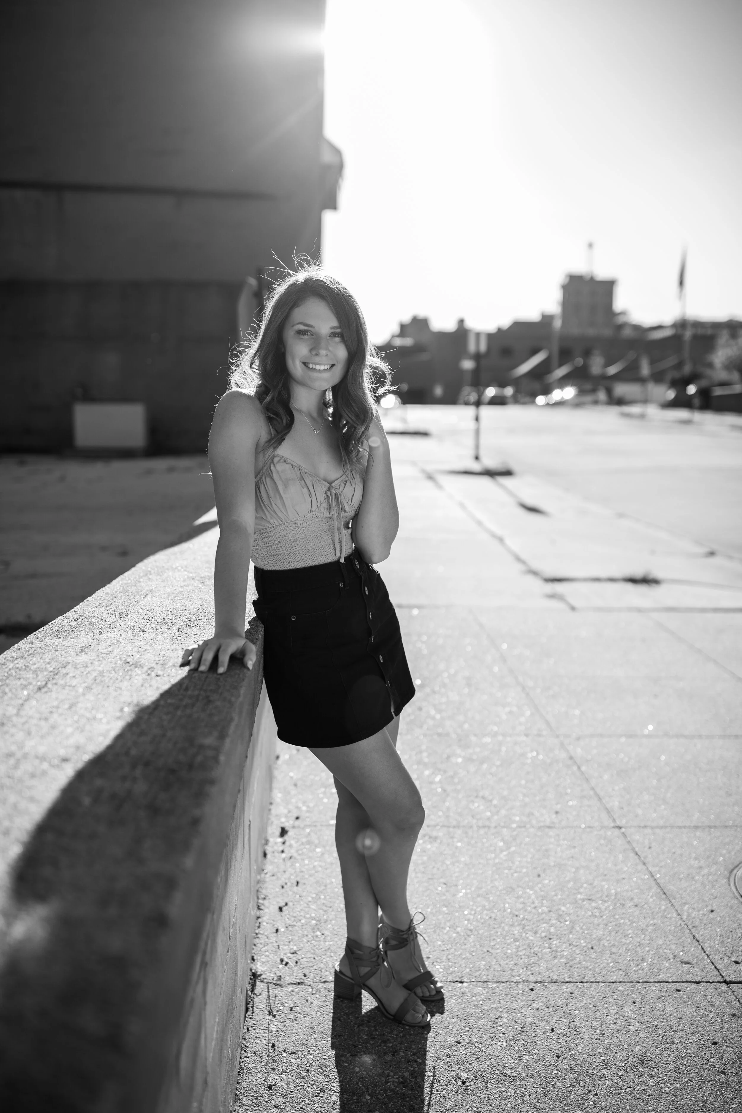 A woman standing outdoors near a concrete ledge, smiling at the camera, with buildings and a clear sky in the background. Black and white photo with bright sunlight.