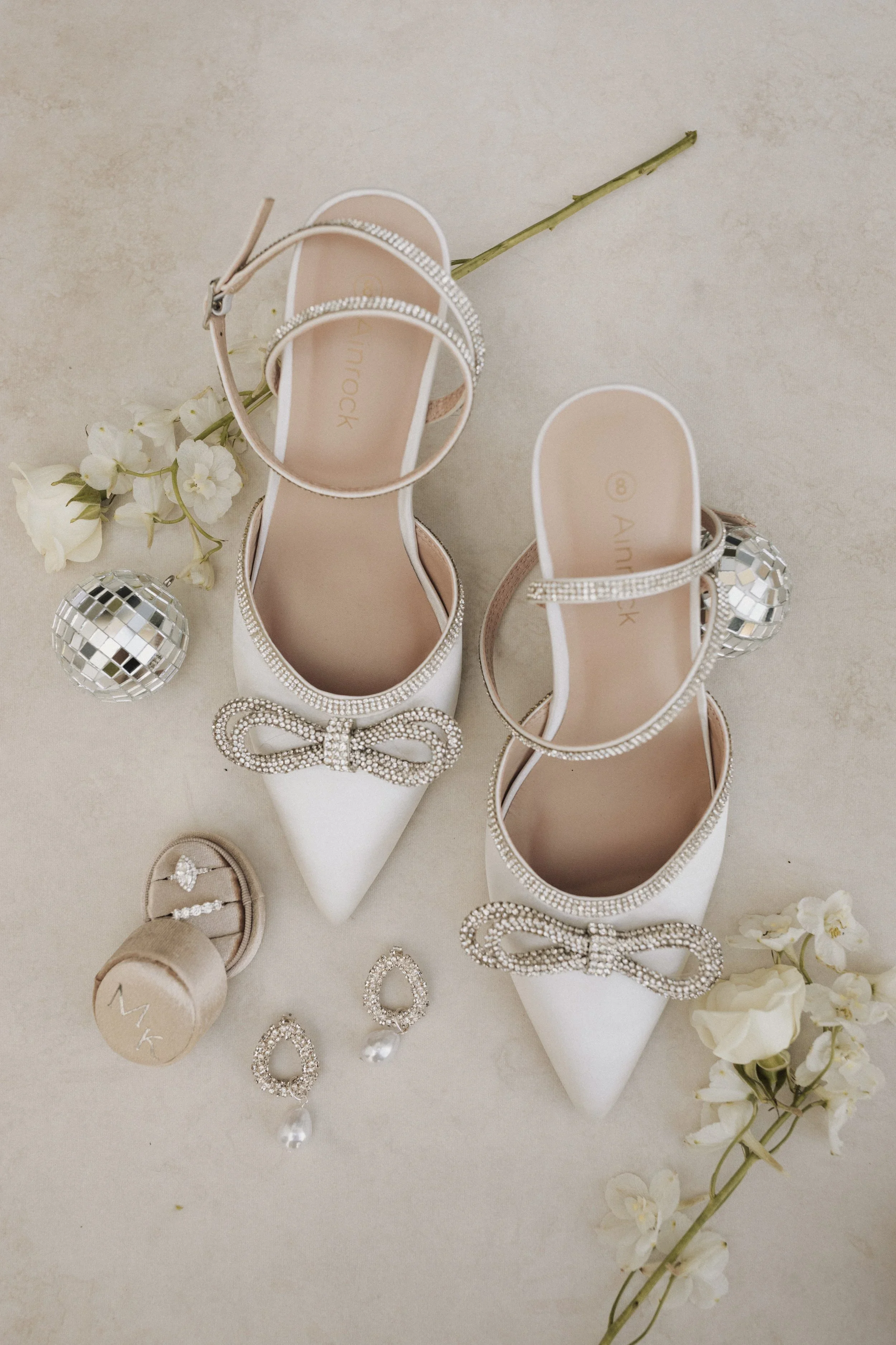 A pair of elegant white high heels with rhinestone details, a pair of pearl earrings, a ring, and a champagne-colored box with the brand initials. The shoes are surrounded by white flowers and a disco ball ornament.