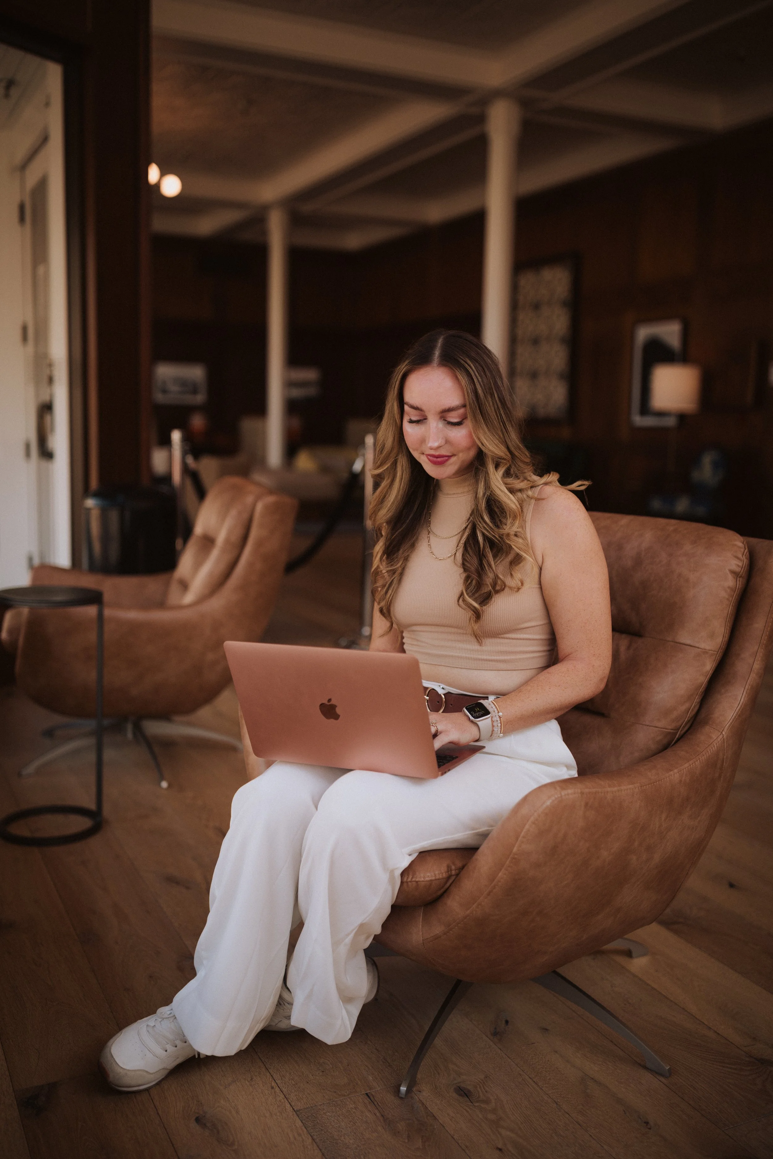 A young woman with long, wavy hair, wearing a beige sleeveless top and white pants, sitting on a tan leather chair, working on a pink laptop in a cozy, wood-paneled room.